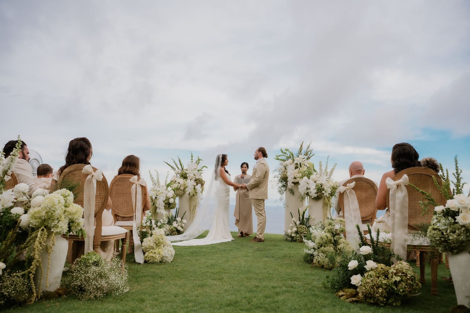 Wedding ceremony on green lawn with white flowers, rattan chairs, and ocean horizon view.