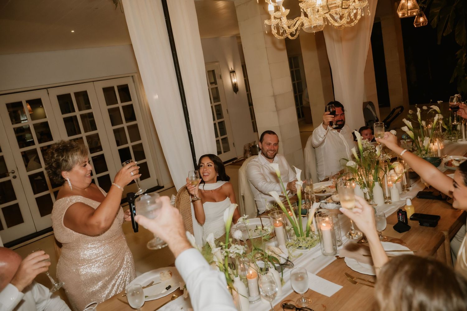 Bride and groom enjoy wedding reception toast with guests raising glasses at candlelit table.