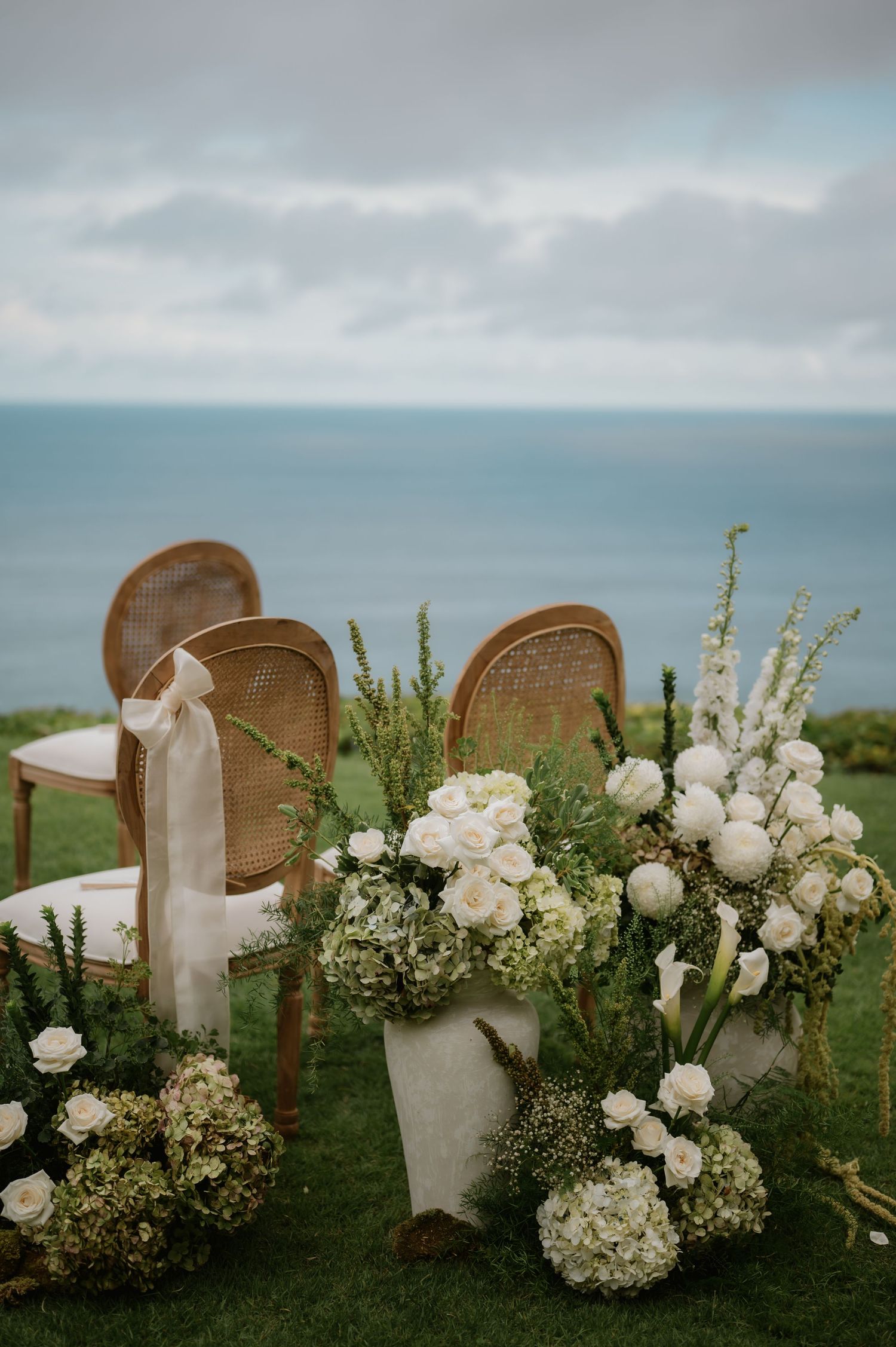 Elegant outdoor wedding ceremony setup with white floral arrangements and rattan chairs overlooking the ocean.