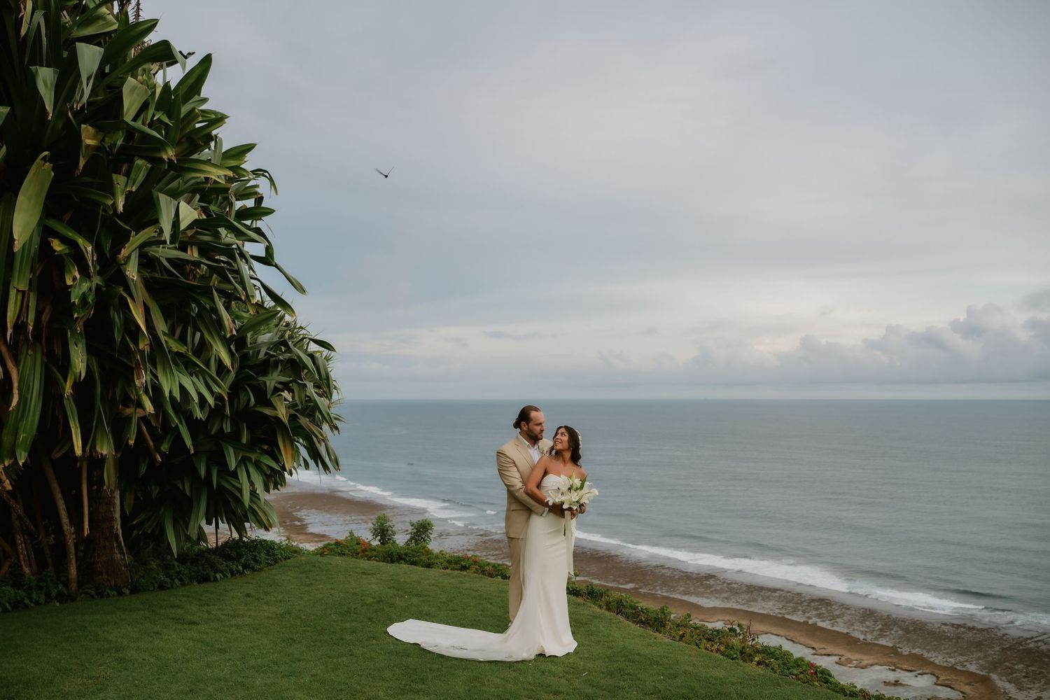Bride and groom embrace on lush green cliff overlooking dramatic ocean coastline at sunset wedding.