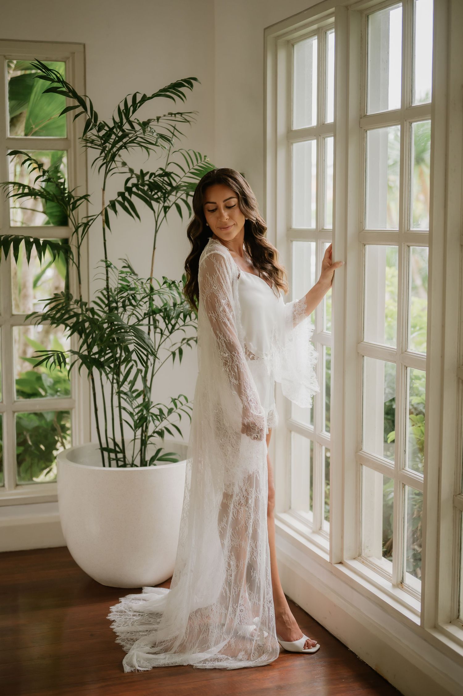 Bride in elegant lace bridal robe stands by sunlit French windows beside a lush indoor palm plant.