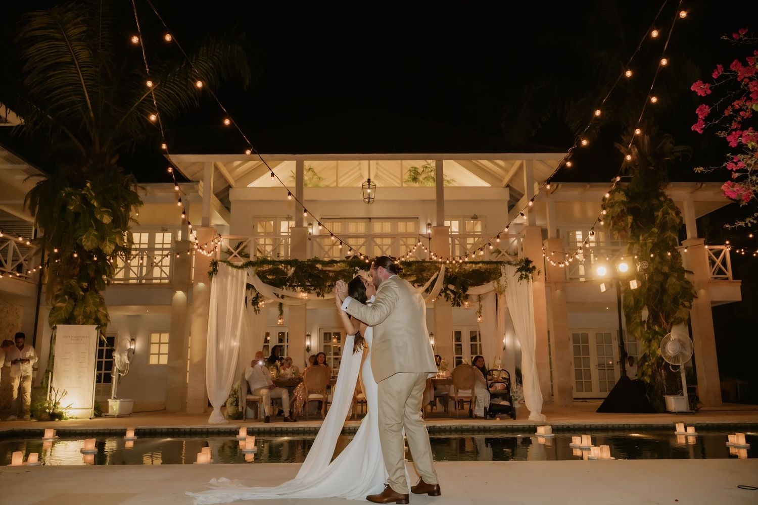 Bride and groom share first dance under string lights beside illuminated pool at night.