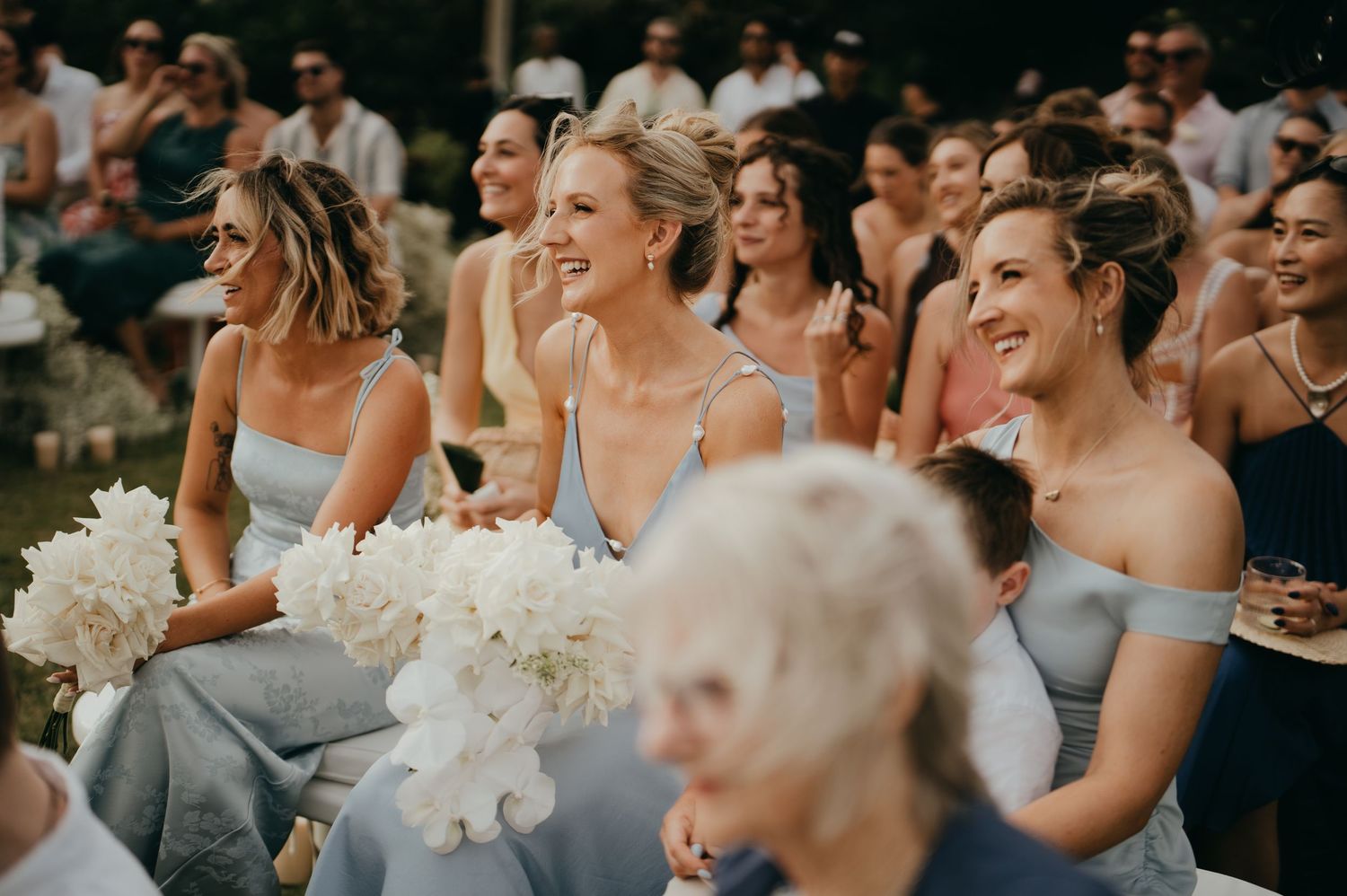 Smiling bridesmaids in light blue off-shoulder dresses holding white peony bouquets at an outdoor wedding ceremony.