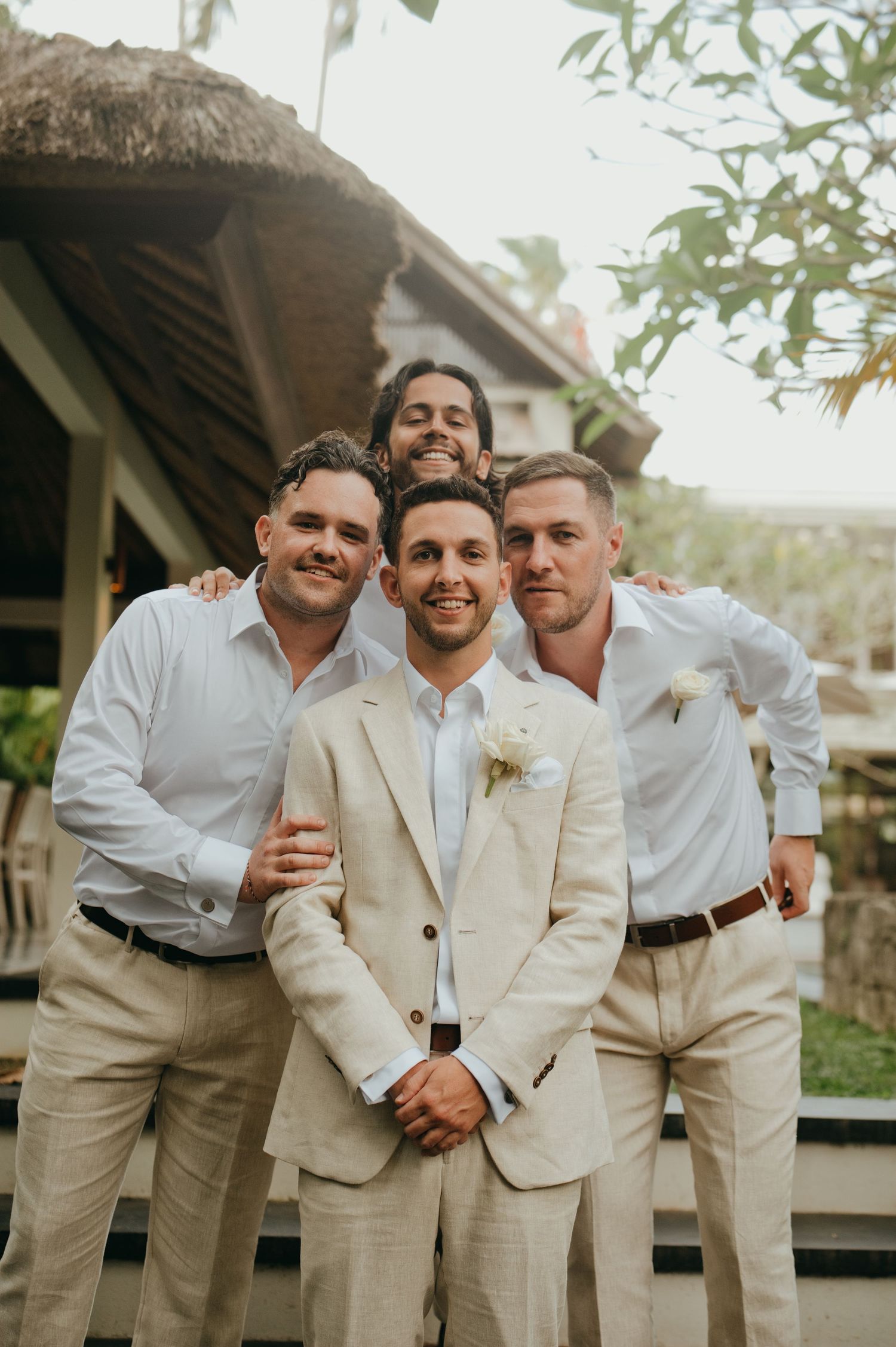 Groom in beige linen suit poses with three groomsmen in white shirts outdoors.