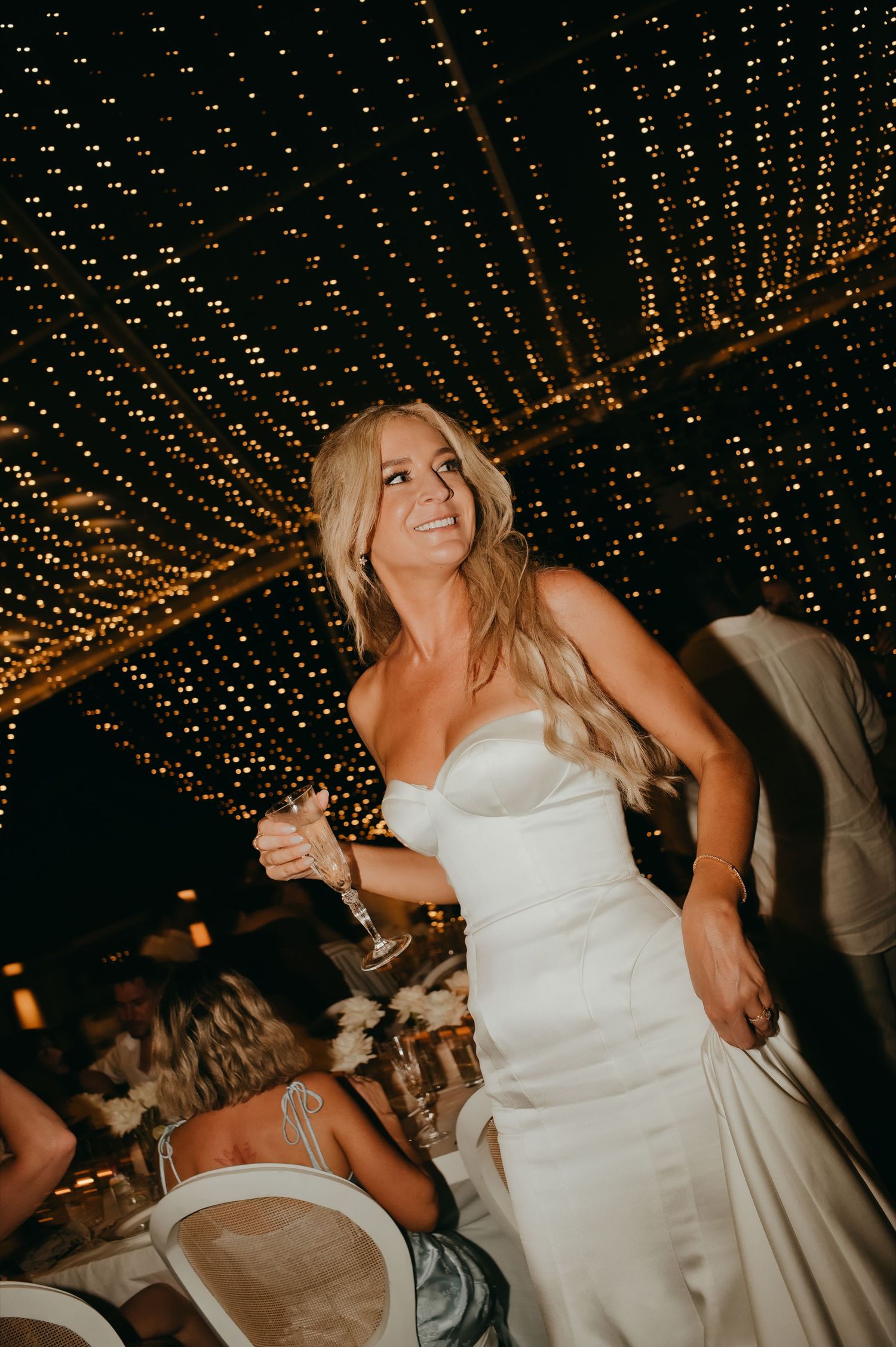 Bride in strapless white gown holding champagne glass under canopy of fairy lights at wedding reception.