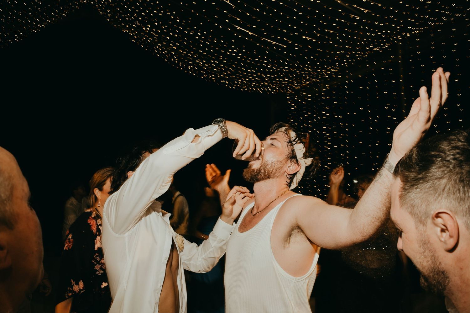 Wedding guests celebrate and dance energetically under a canopy of twinkling fairy lights at the reception.