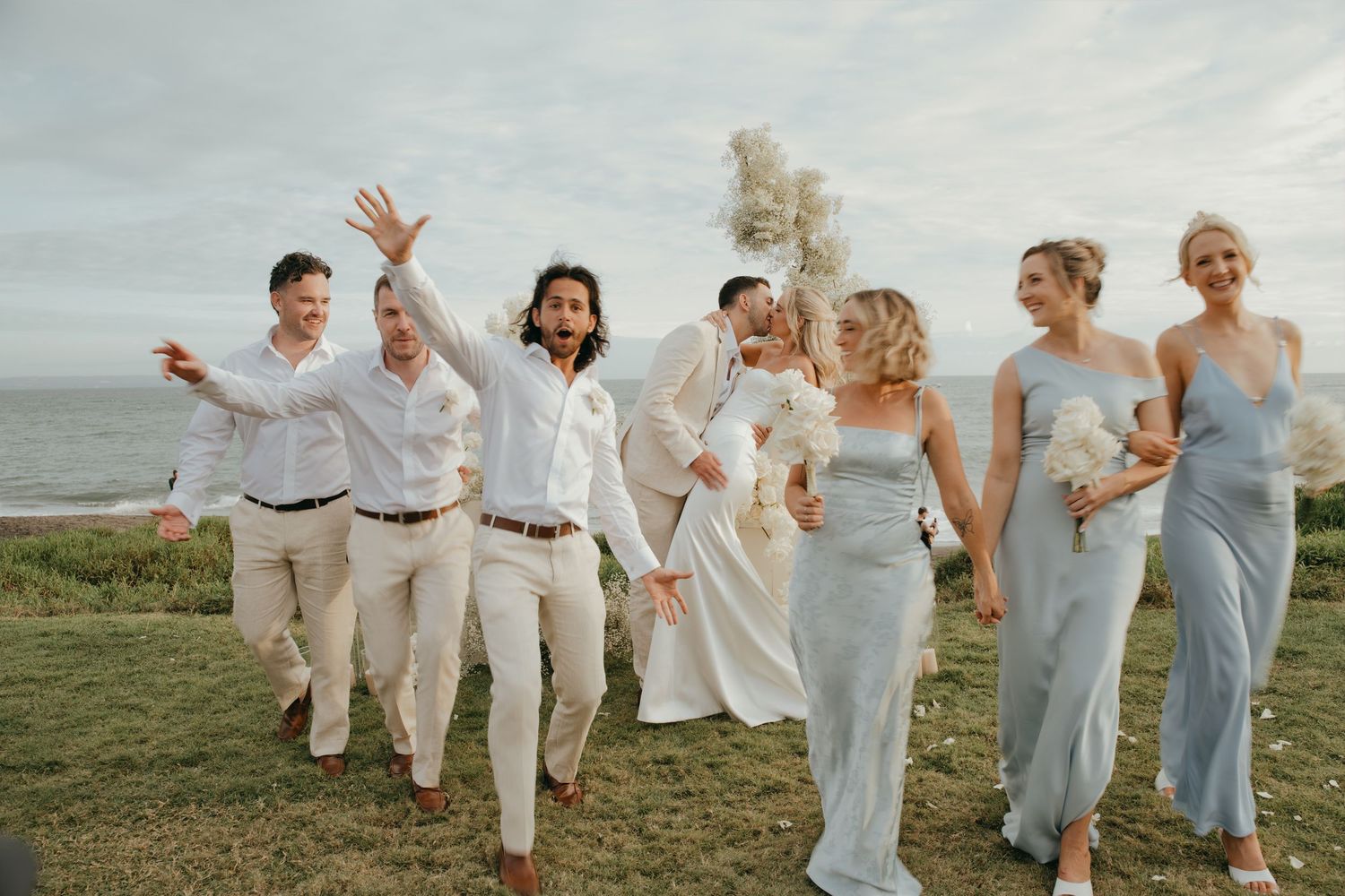 Wedding party celebrates on coastal lawn with bridesmaids in blue dresses and groomsmen in white shirts.