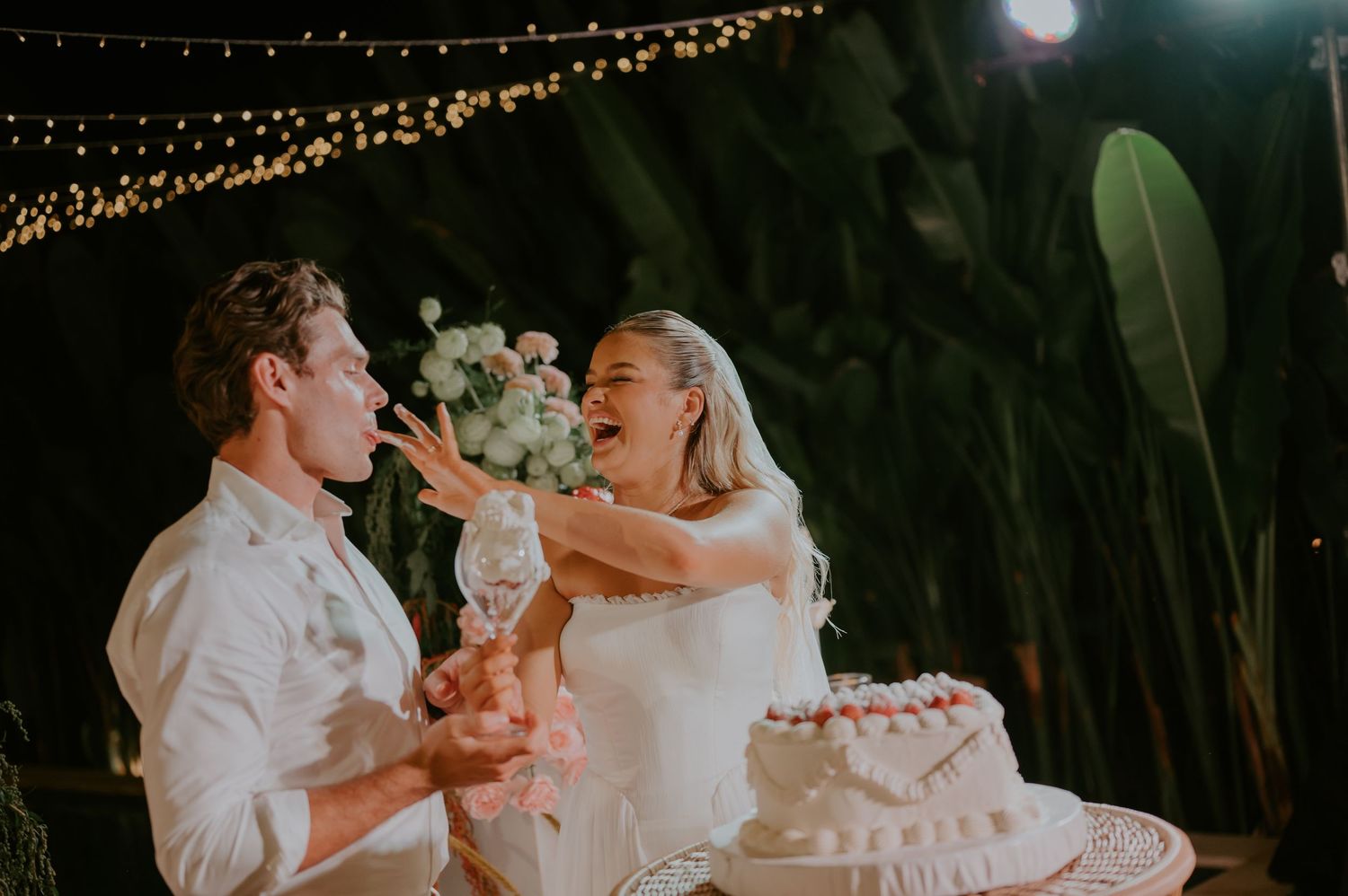 Bride and groom laughing as they smash wedding cake into each other's faces at their outdoor night reception.