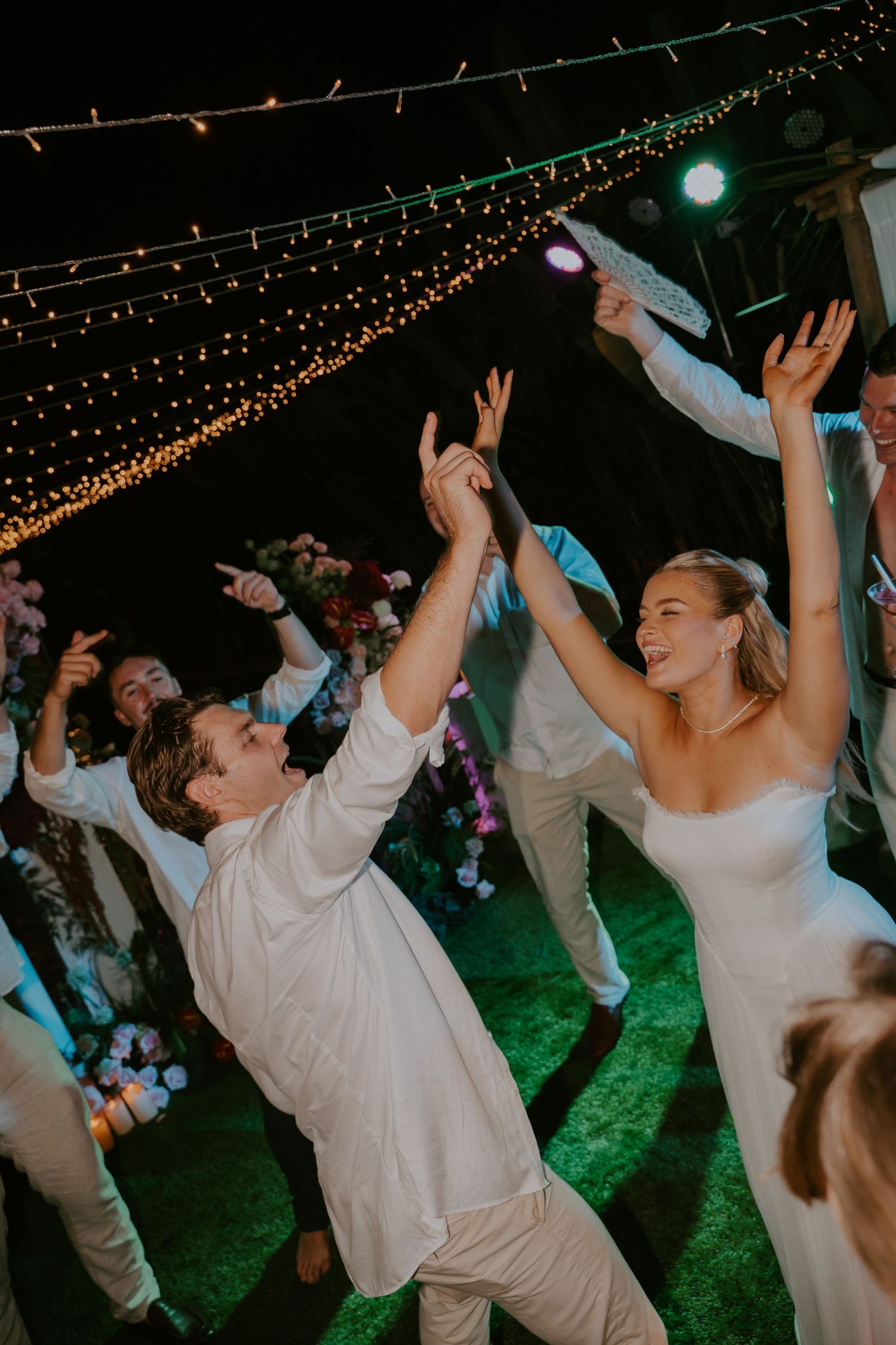 Bride and groom dance joyfully with raised hands under string lights at their outdoor wedding reception.