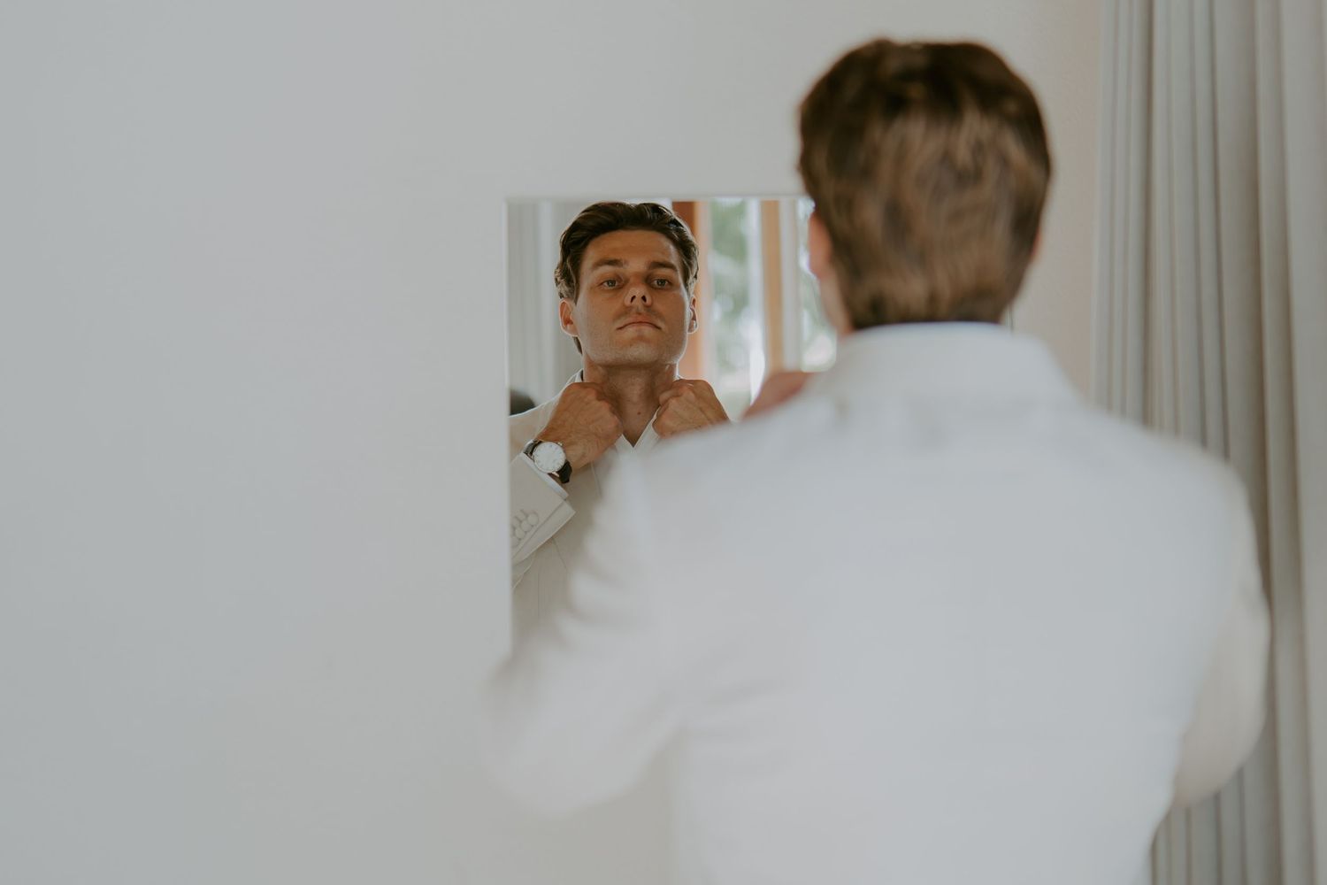 Groom in white shirt adjusting his collar while looking in a mirror during wedding preparations.