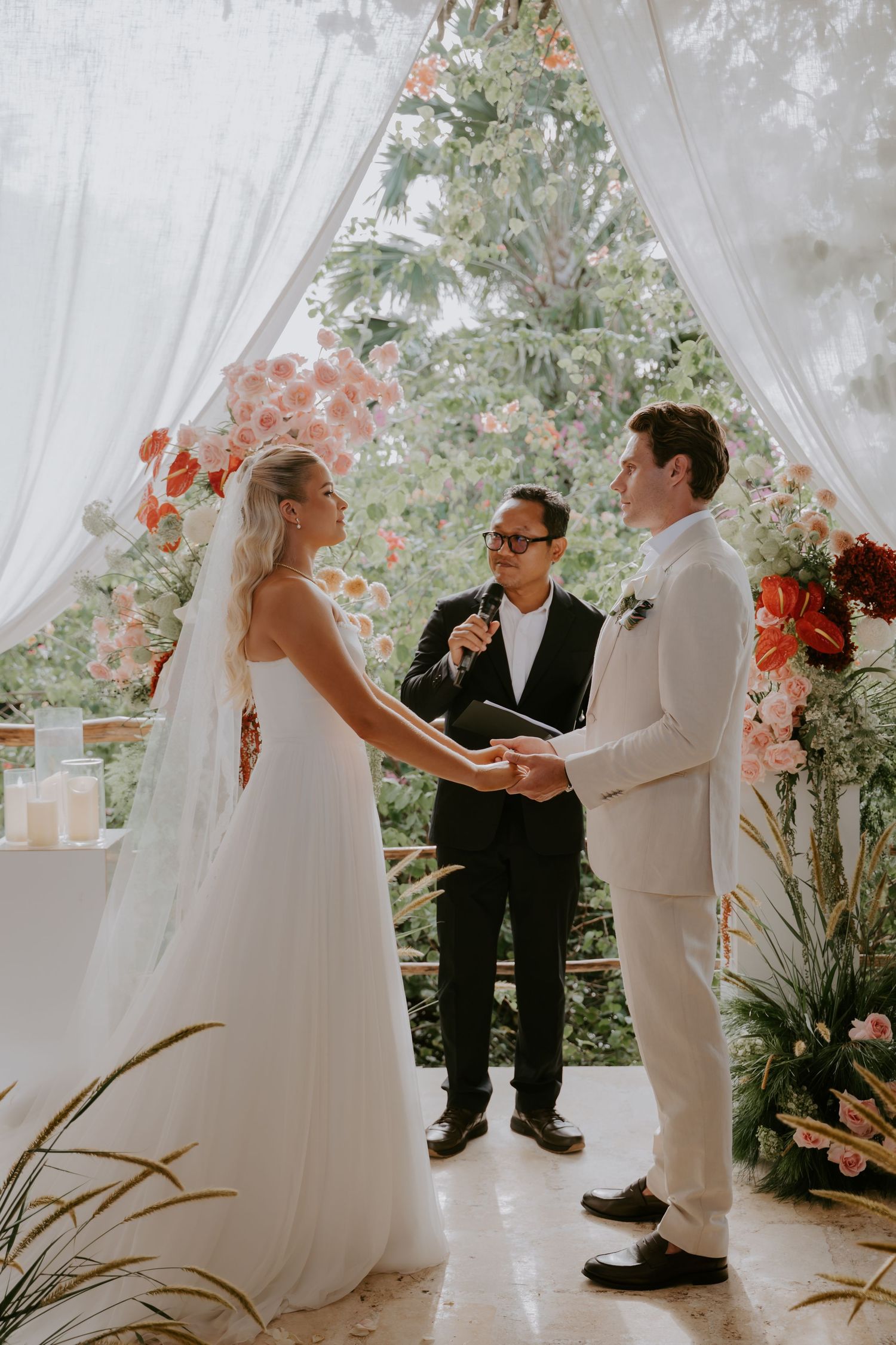 Bride and groom exchange vows beneath floral arch with officiant at tropical outdoor wedding ceremony.
