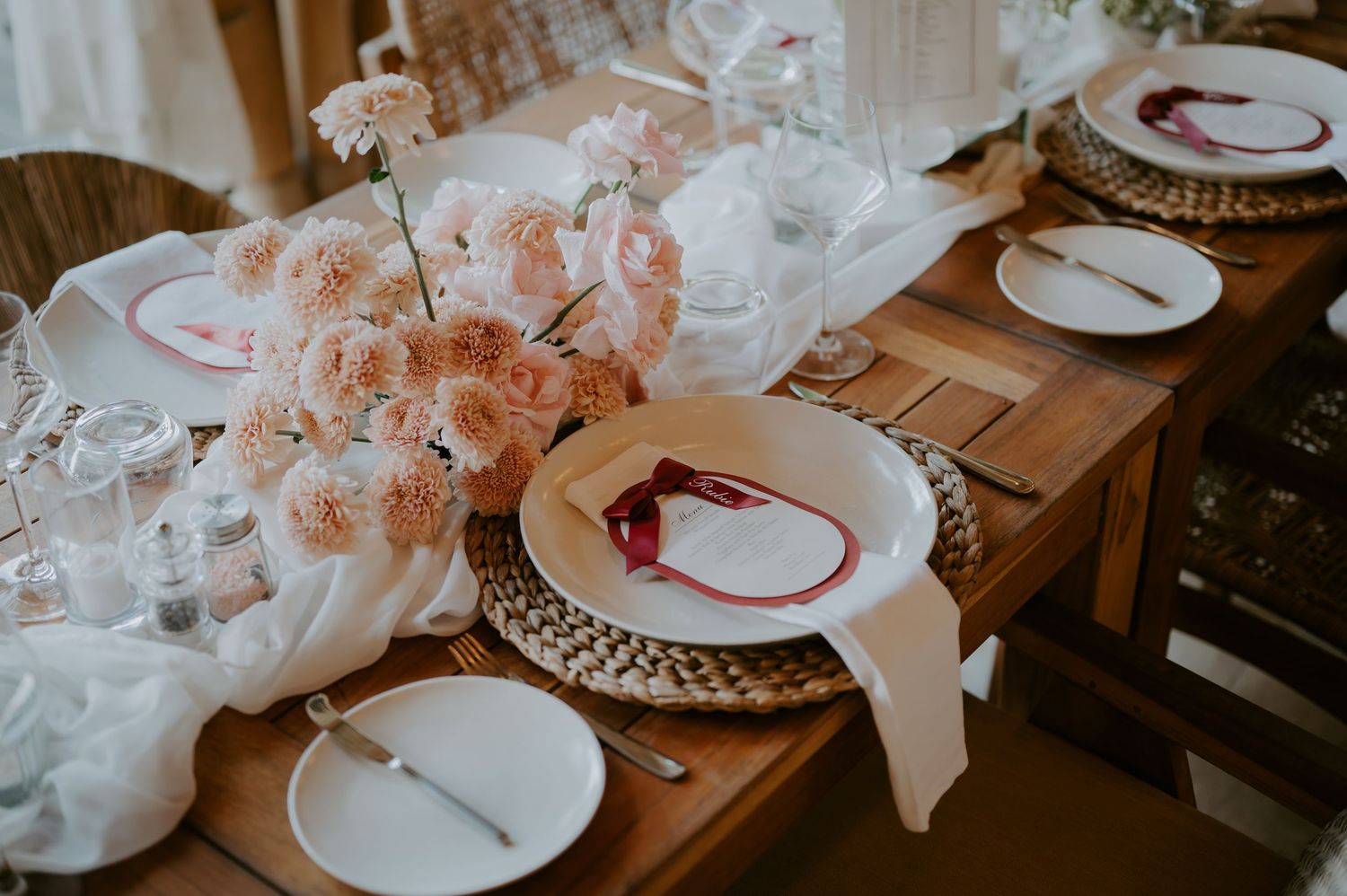 Elegant wedding reception table set with blush carnations, white plates, wicker chargers, and wine glasses.
