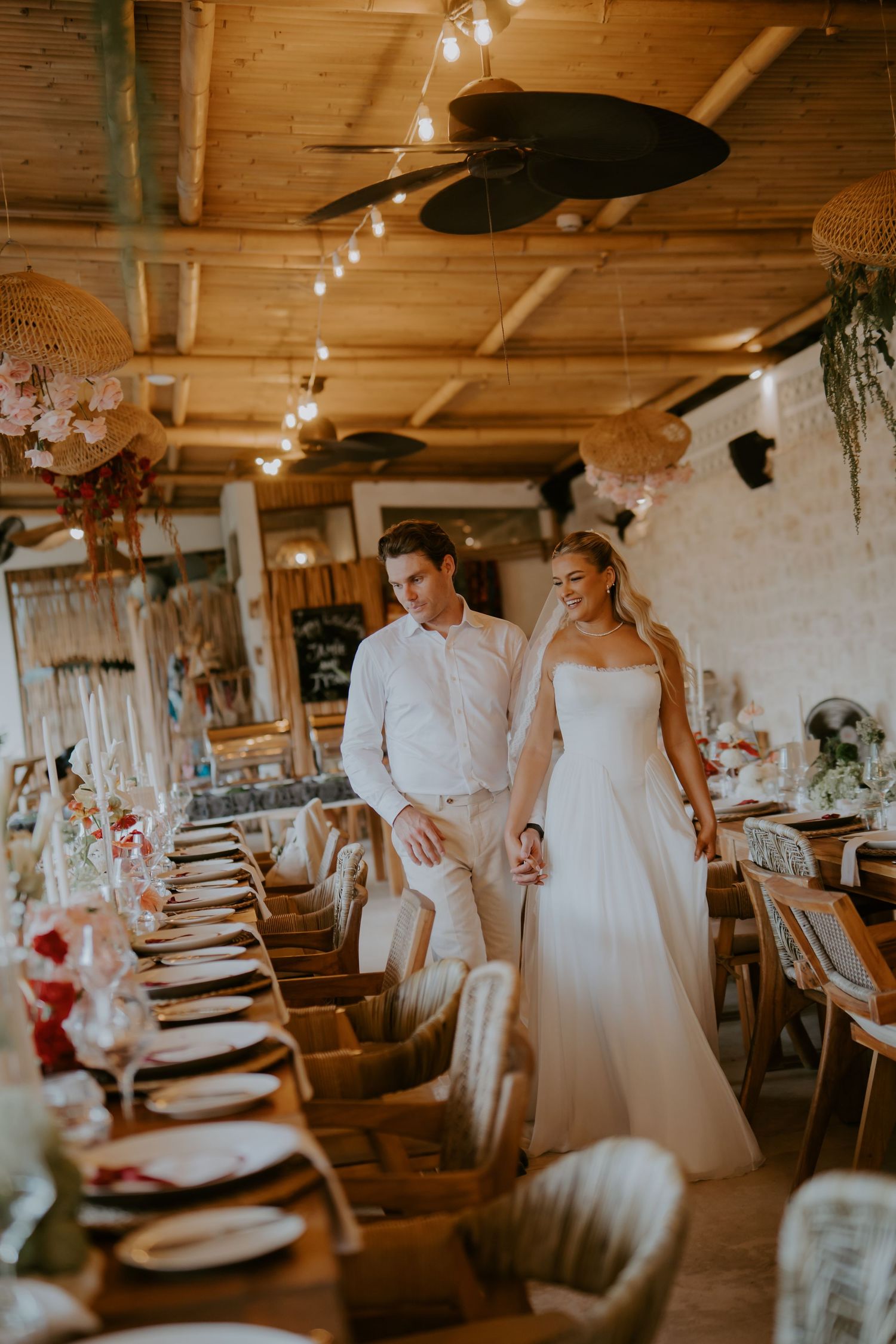 Bride and groom walking hand in hand through a boho wedding reception venue with string lights and long tables.