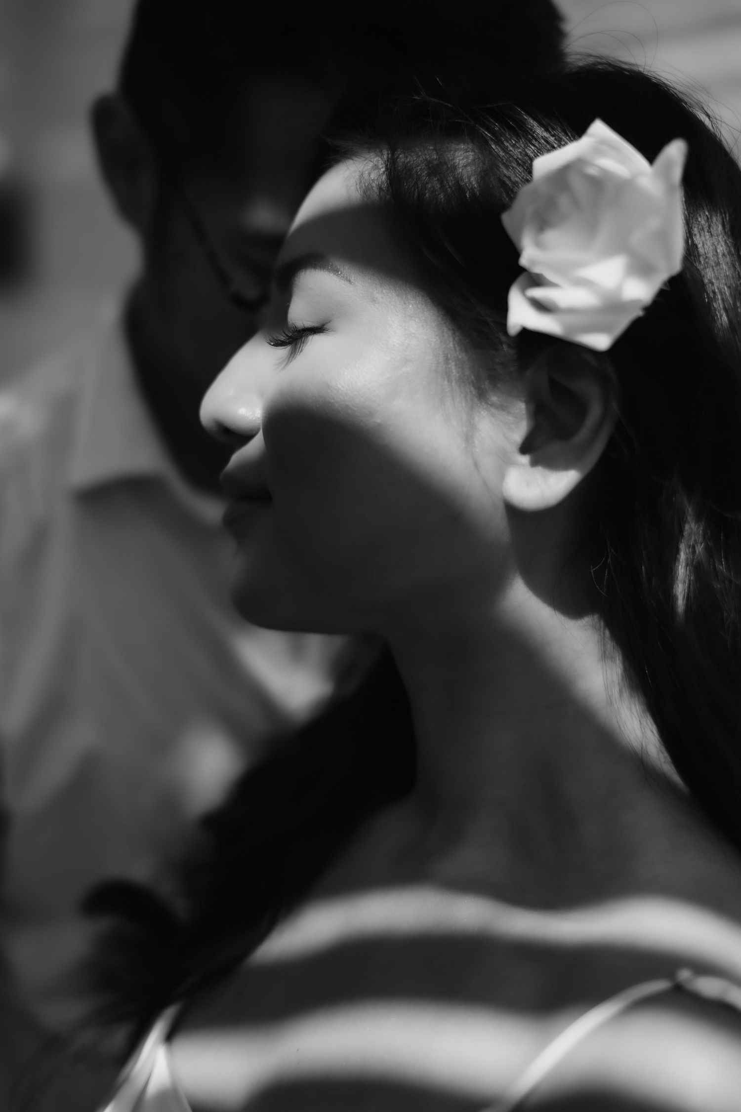 Black and white portrait of a young woman with a flower in her hair, eyes closed, standing before a man.
