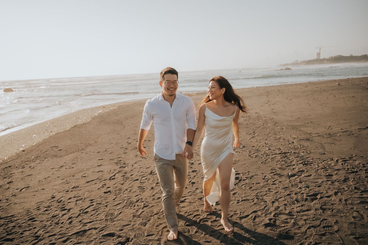 Happy couple holding hands and running on a sandy beach at golden hour with ocean waves in the background.