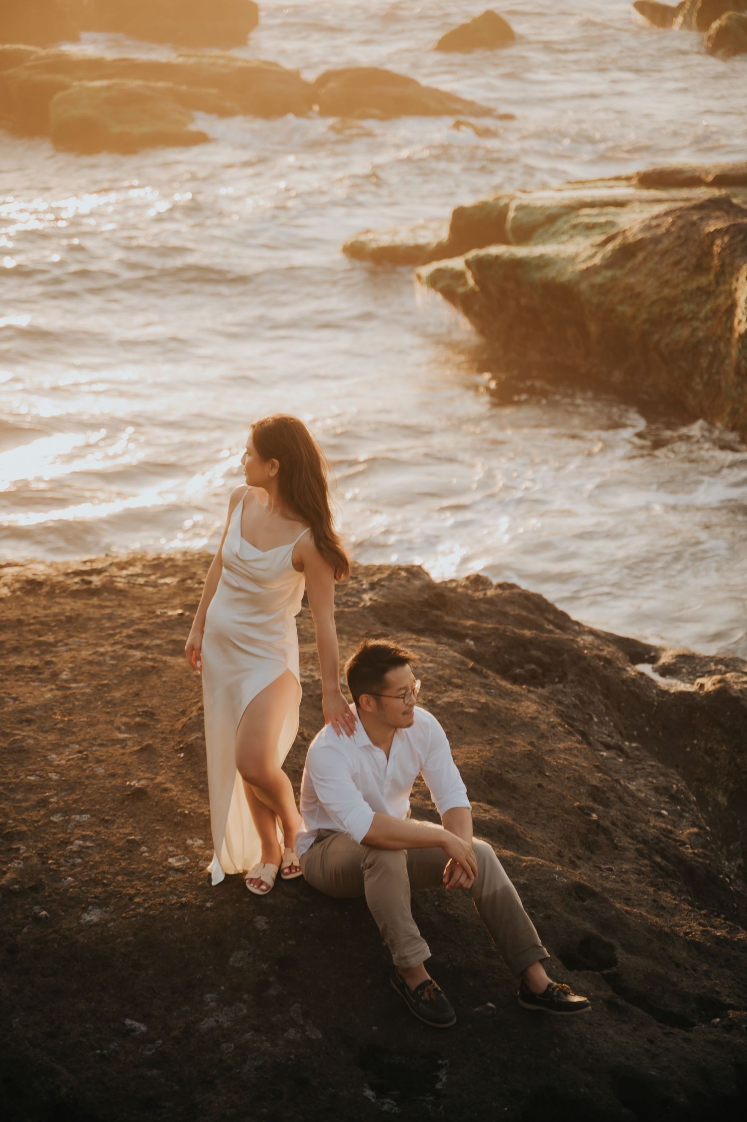 Couple in white attire poses on rocky coastal cliffs at golden sunset with ocean waves in background.
