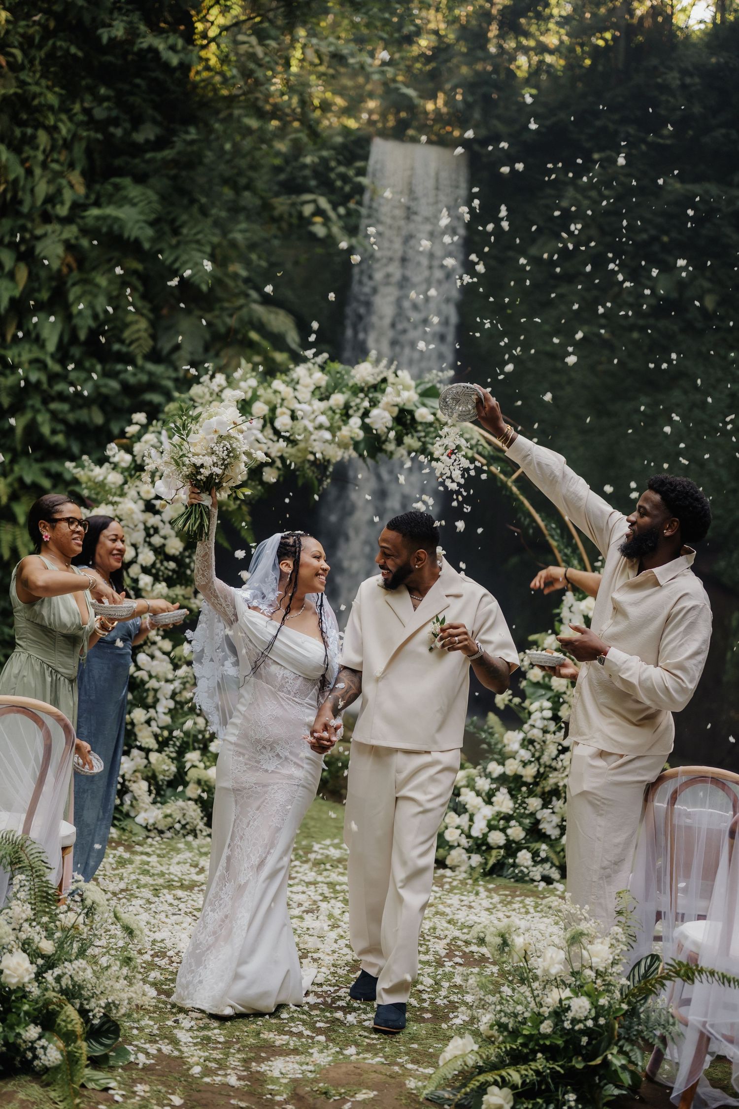 Happy newlyweds celebrate with confetti toss as guests cheer beside a lush tropical waterfall backdrop.