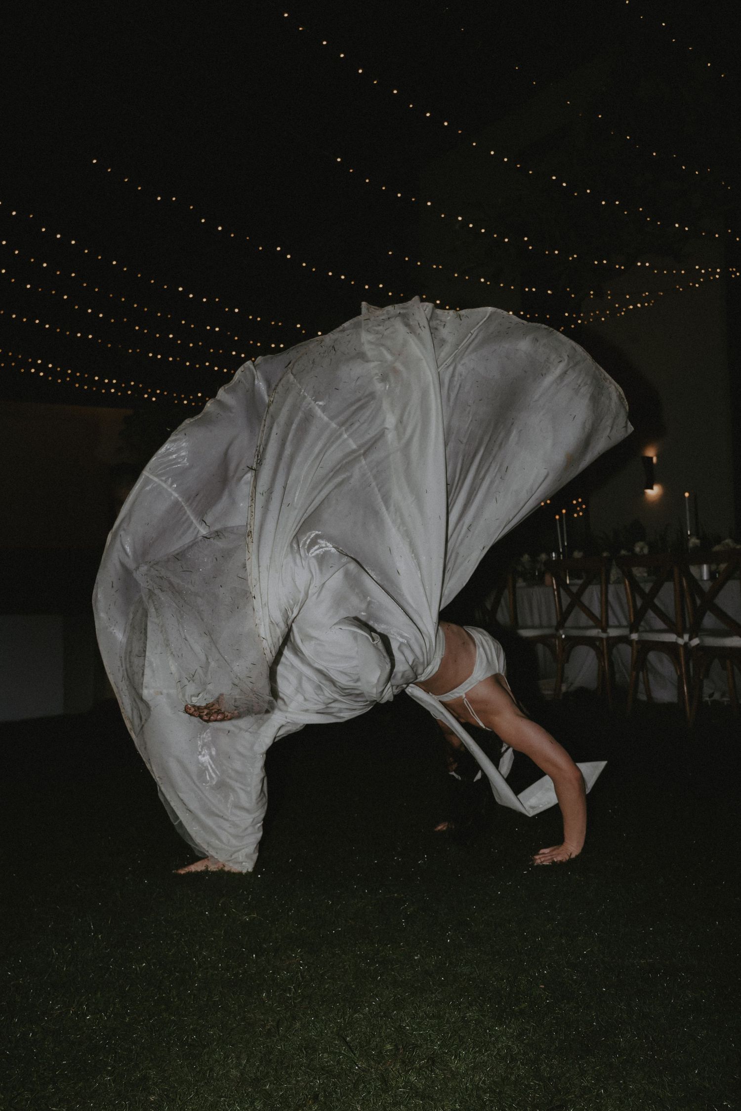 A sequence of photos showing someone in a flowing white dress doing a backflip on grass at night under string lights.