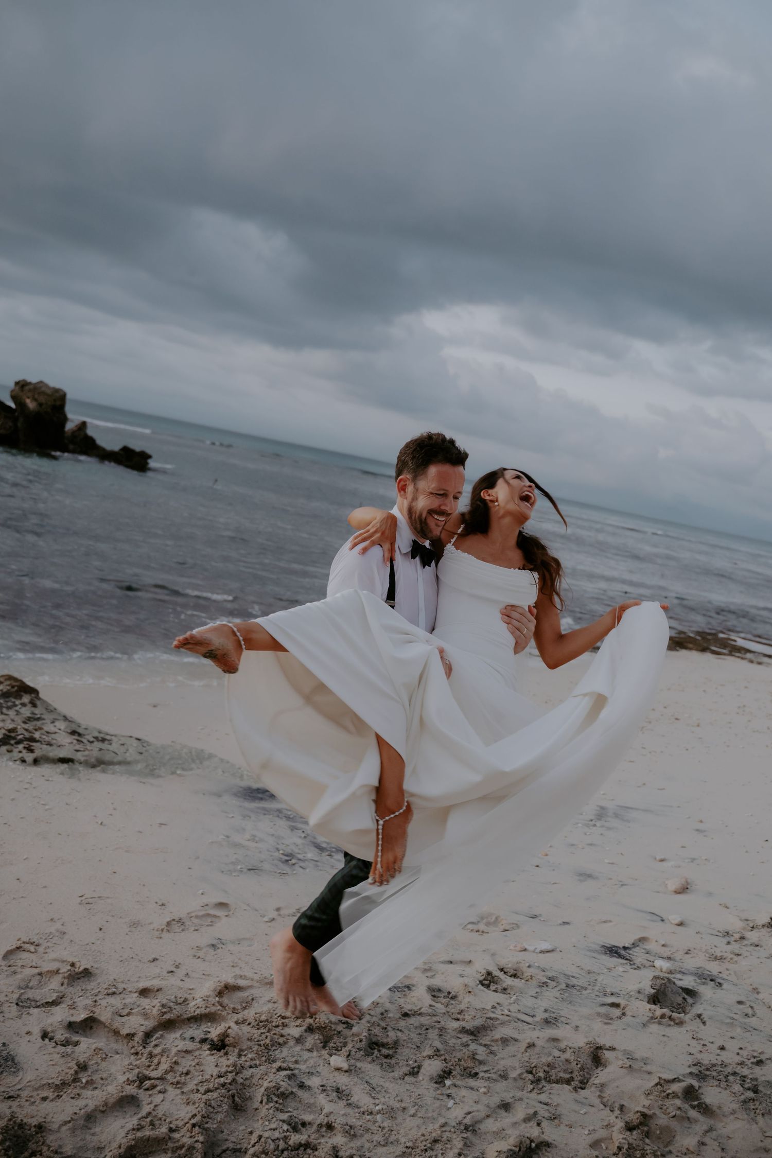 A couple shares a romantic moment on a cloudy beach as the bride's white dress flows dramatically in the wind.