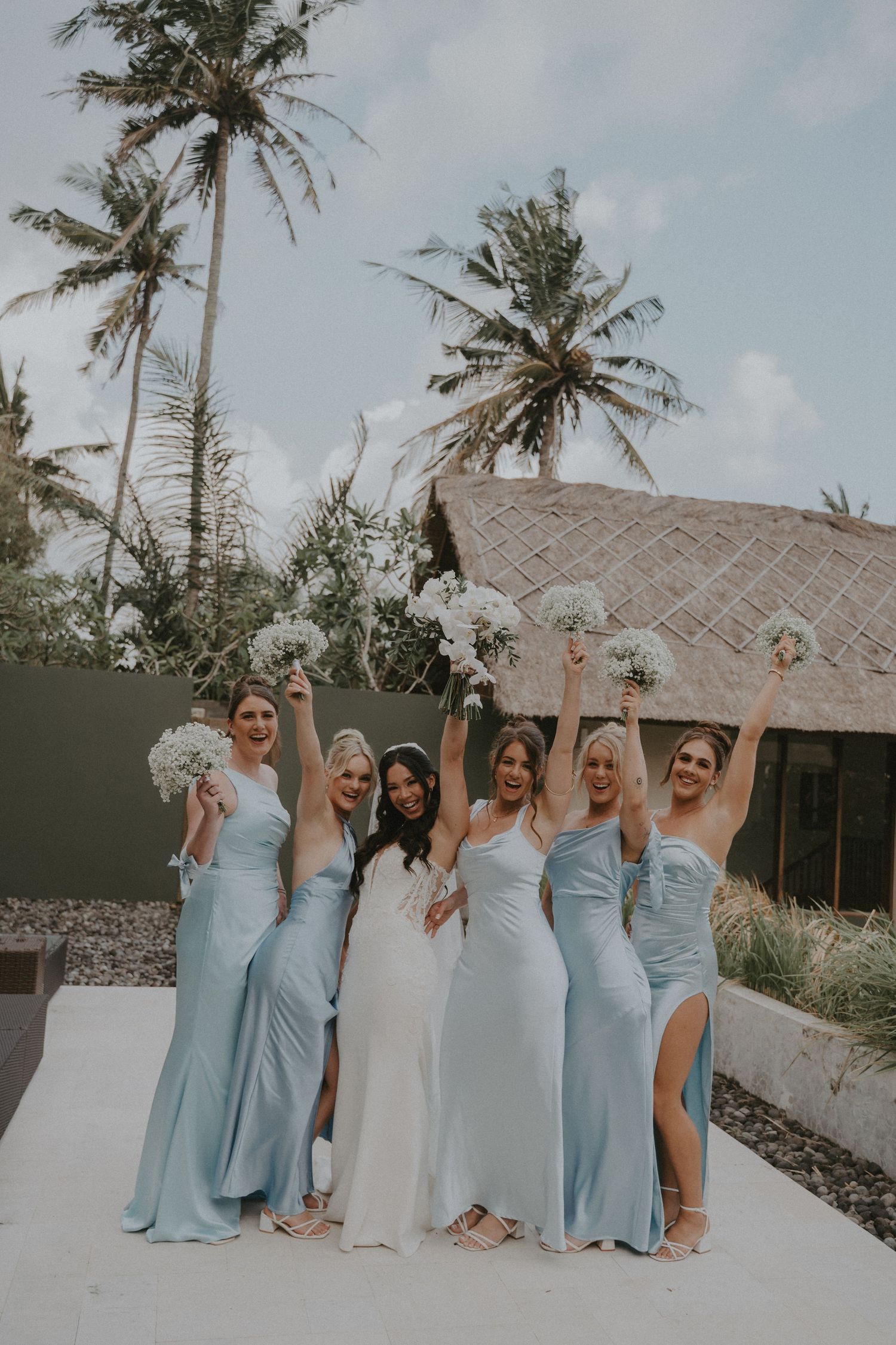 Bridesmaids in light blue dresses pose together with palm trees and a tropical resort building in the background.