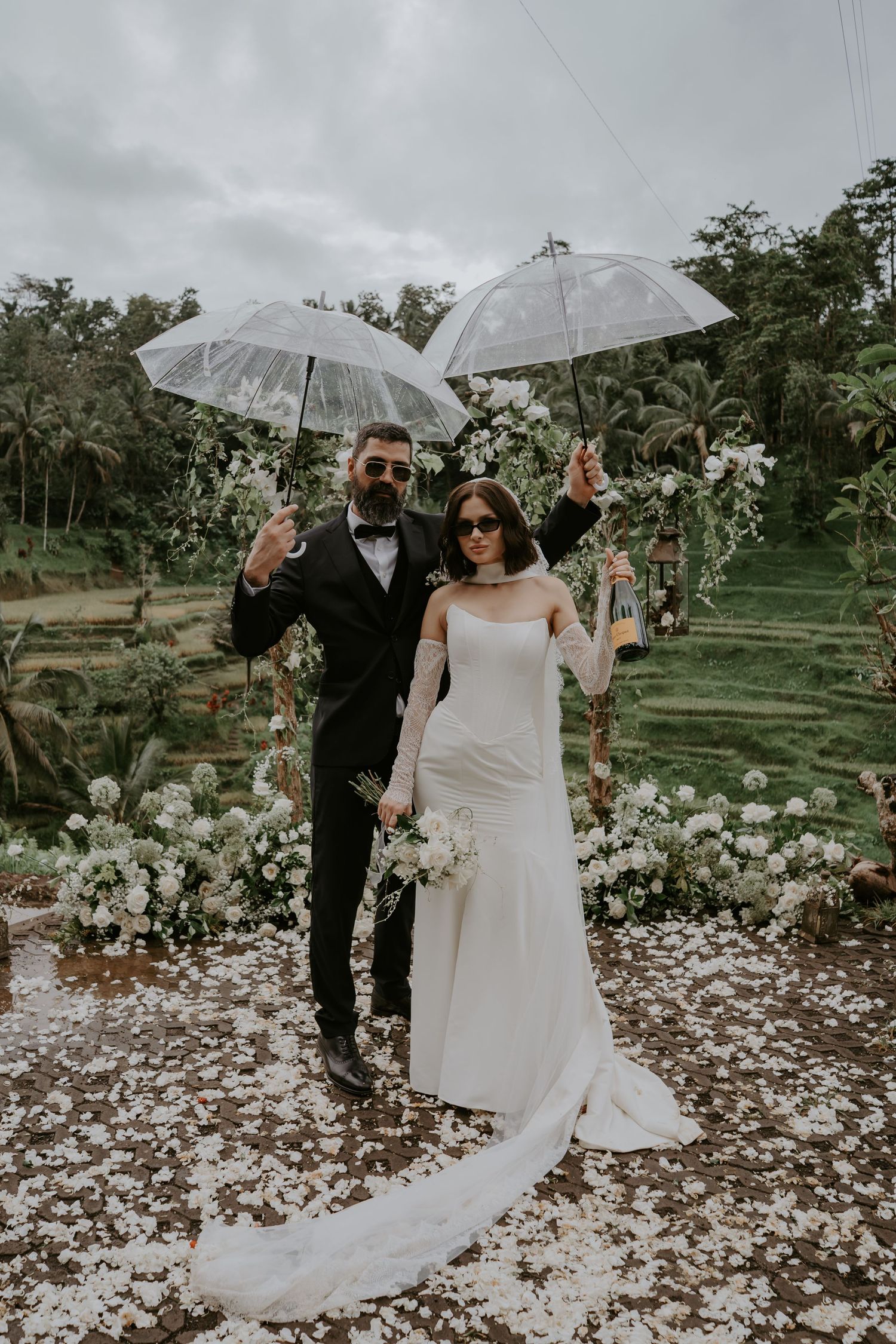 A couple poses with clear umbrellas in a lush garden setting during their rainy day wedding celebration.