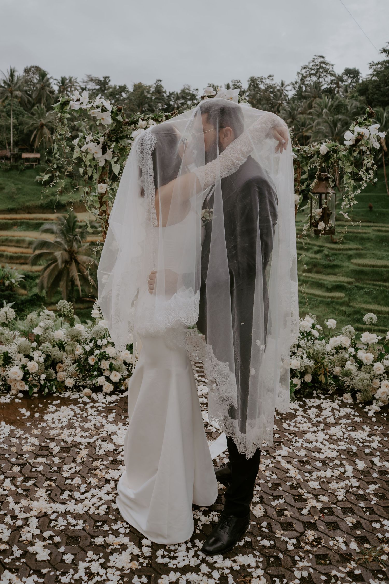 A romantic wedding couple shares intimate moments under a veil in a garden setting with scattered flower petals on the ground.