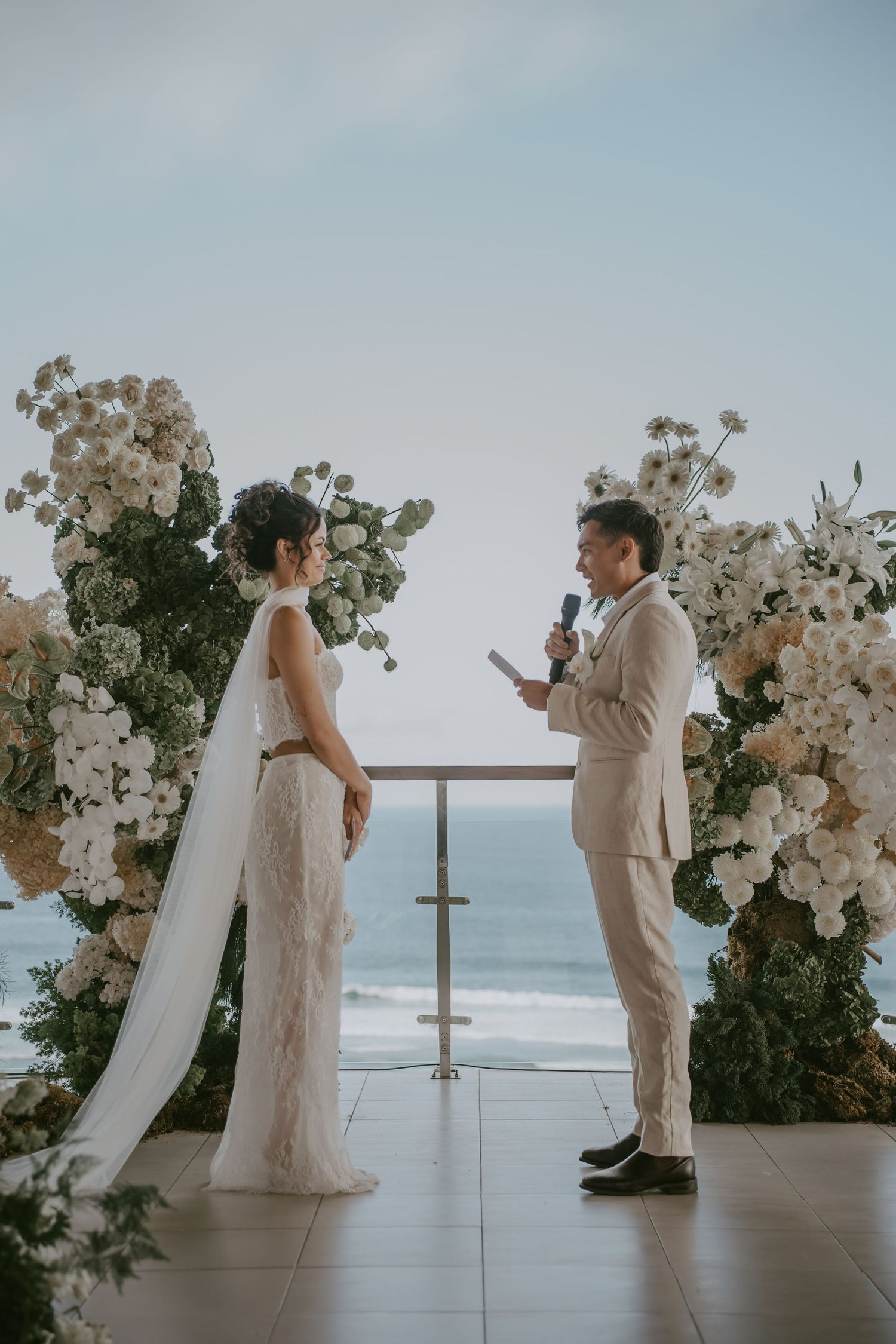 A romantic beach wedding ceremony with white floral arrangements and ocean views as a couple exchanges vows.