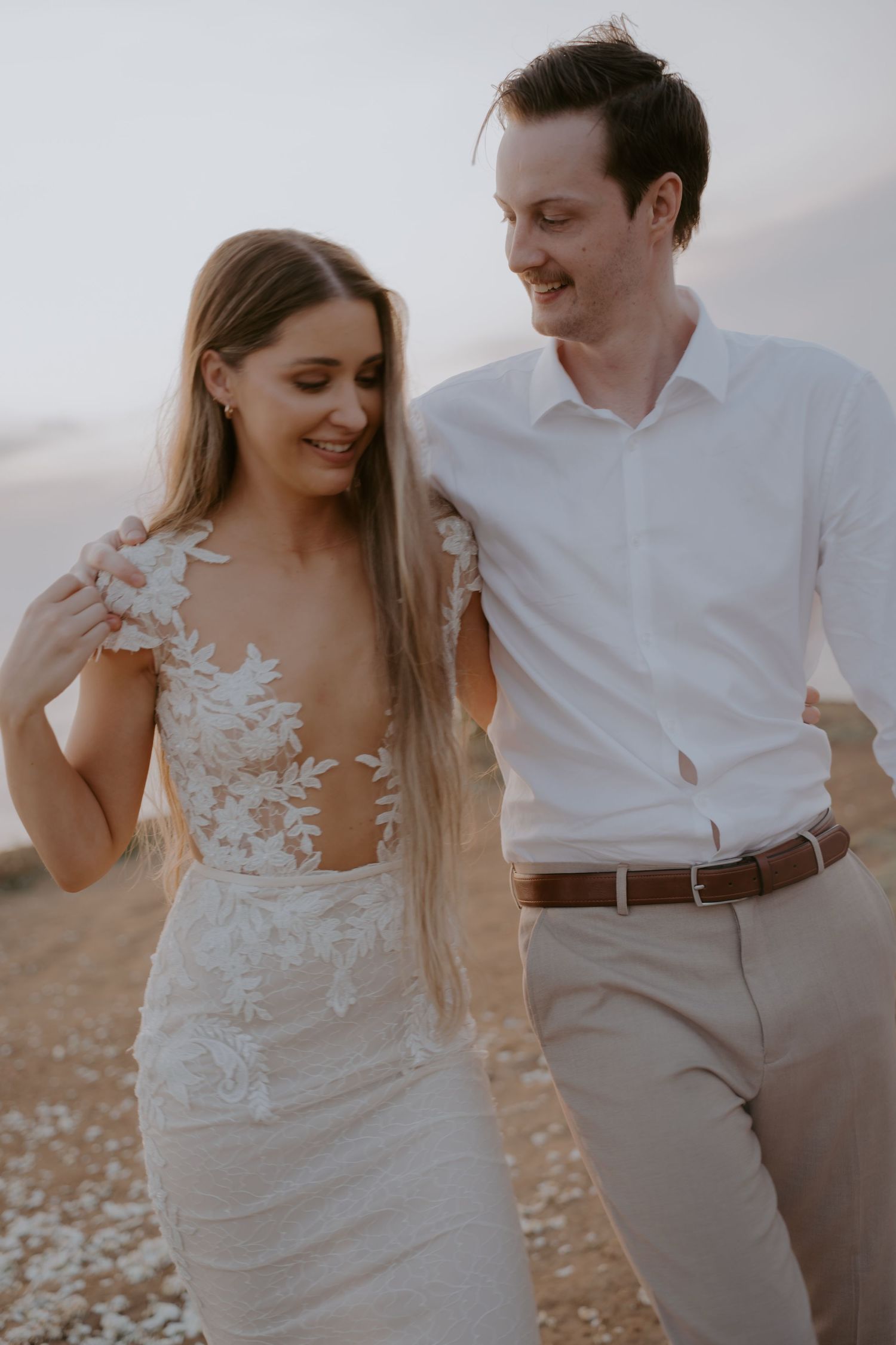 A romantic beach wedding couple laughs and embraces while walking together in flowing white attire at sunset.