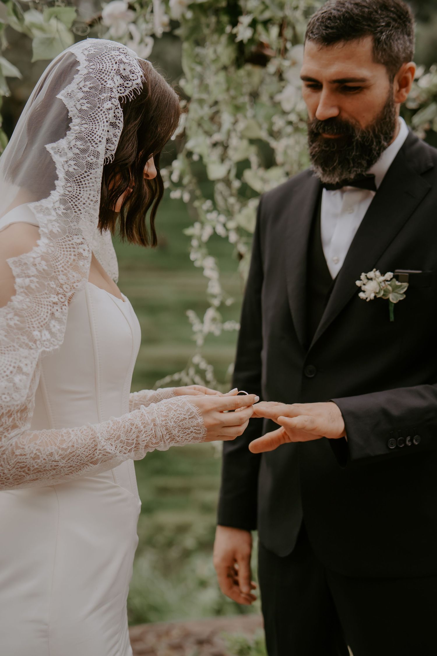 A bride in lace sleeves places a ring on the groom's hand during an outdoor wedding ceremony.