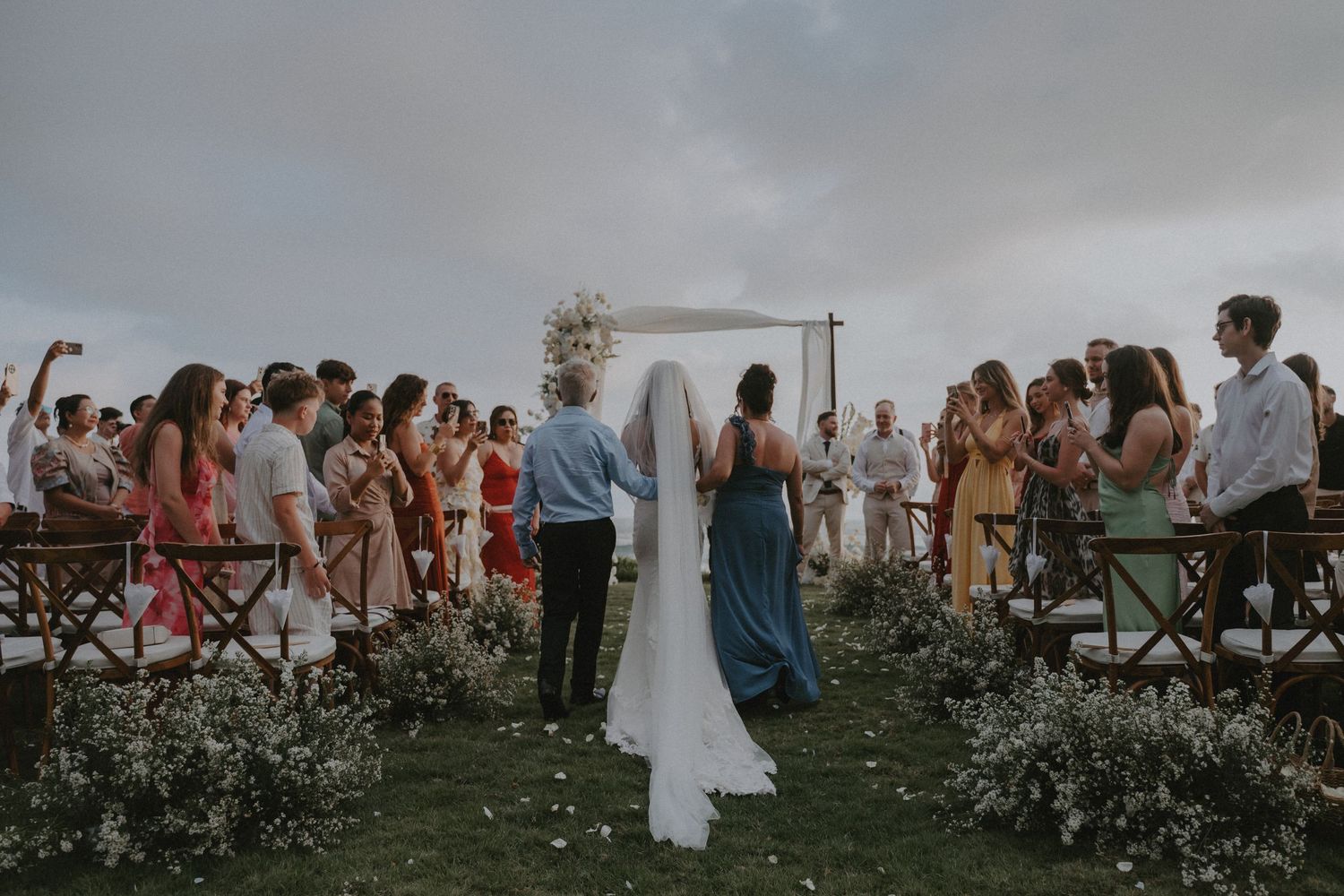 Outdoor wedding ceremony on a cloudy day with guests seated in wooden chairs surrounded by wildflowers and greenery.