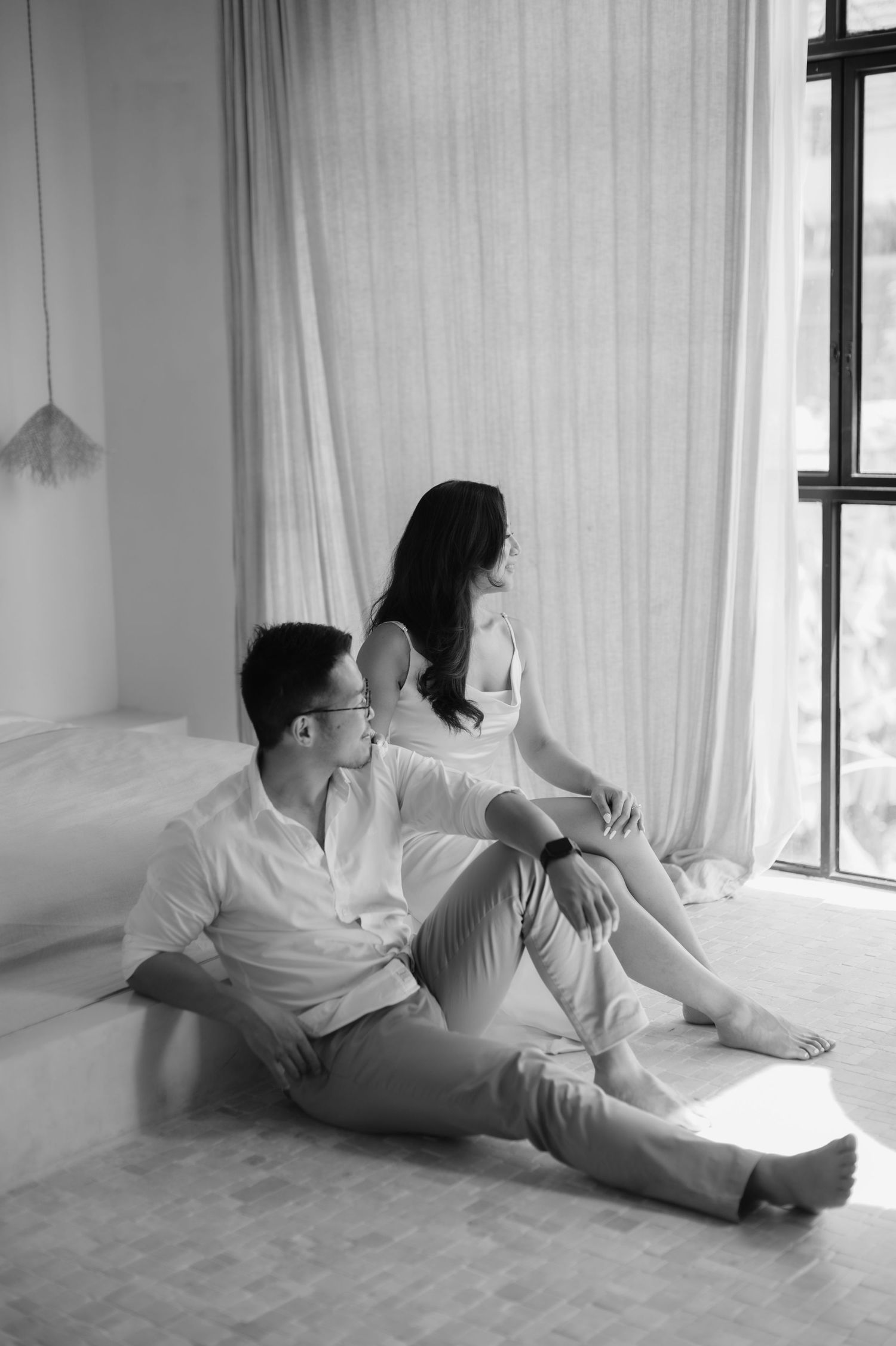 Black and white photo of a couple sitting on a floor near a window, man gazing at woman who looks away.