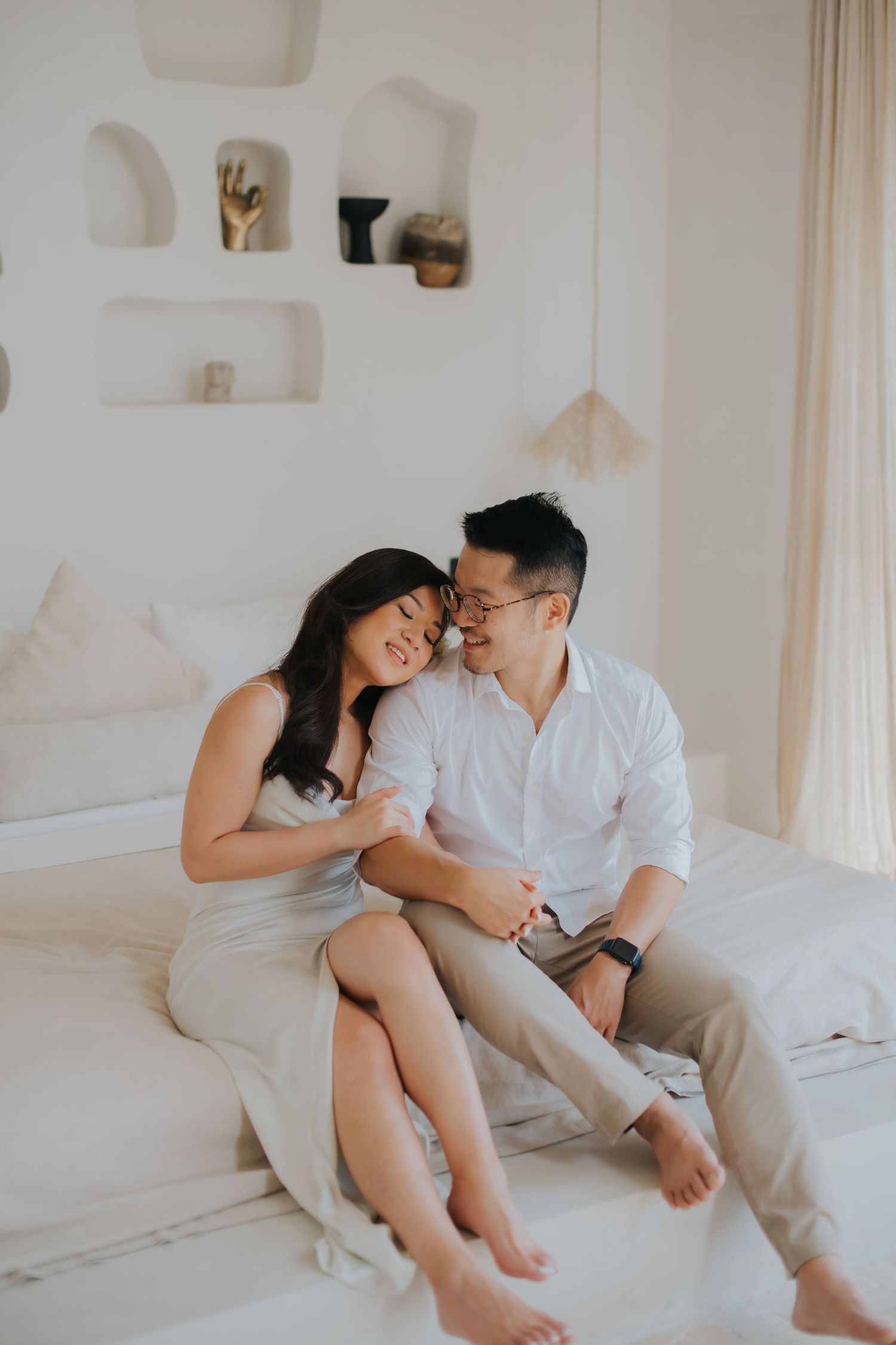 A happy Asian couple sits together on a white bed in a bright, minimalist bedroom, sharing an intimate moment.
