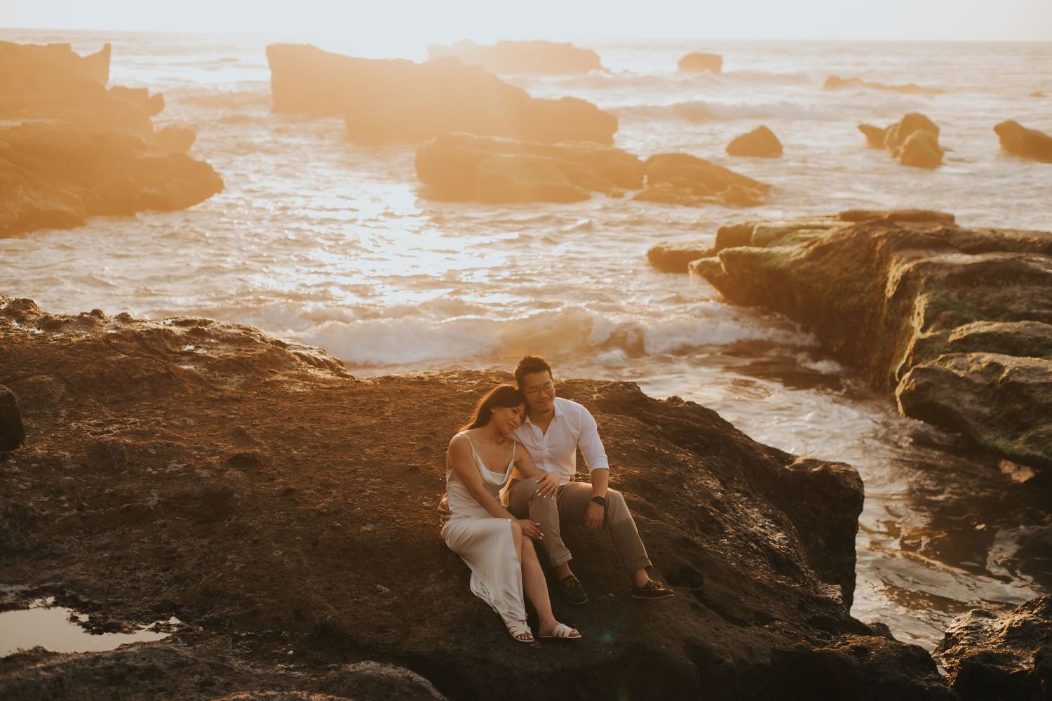 Couple sitting together on rocky coastal cliffs at golden sunset with ocean waves crashing behind them.