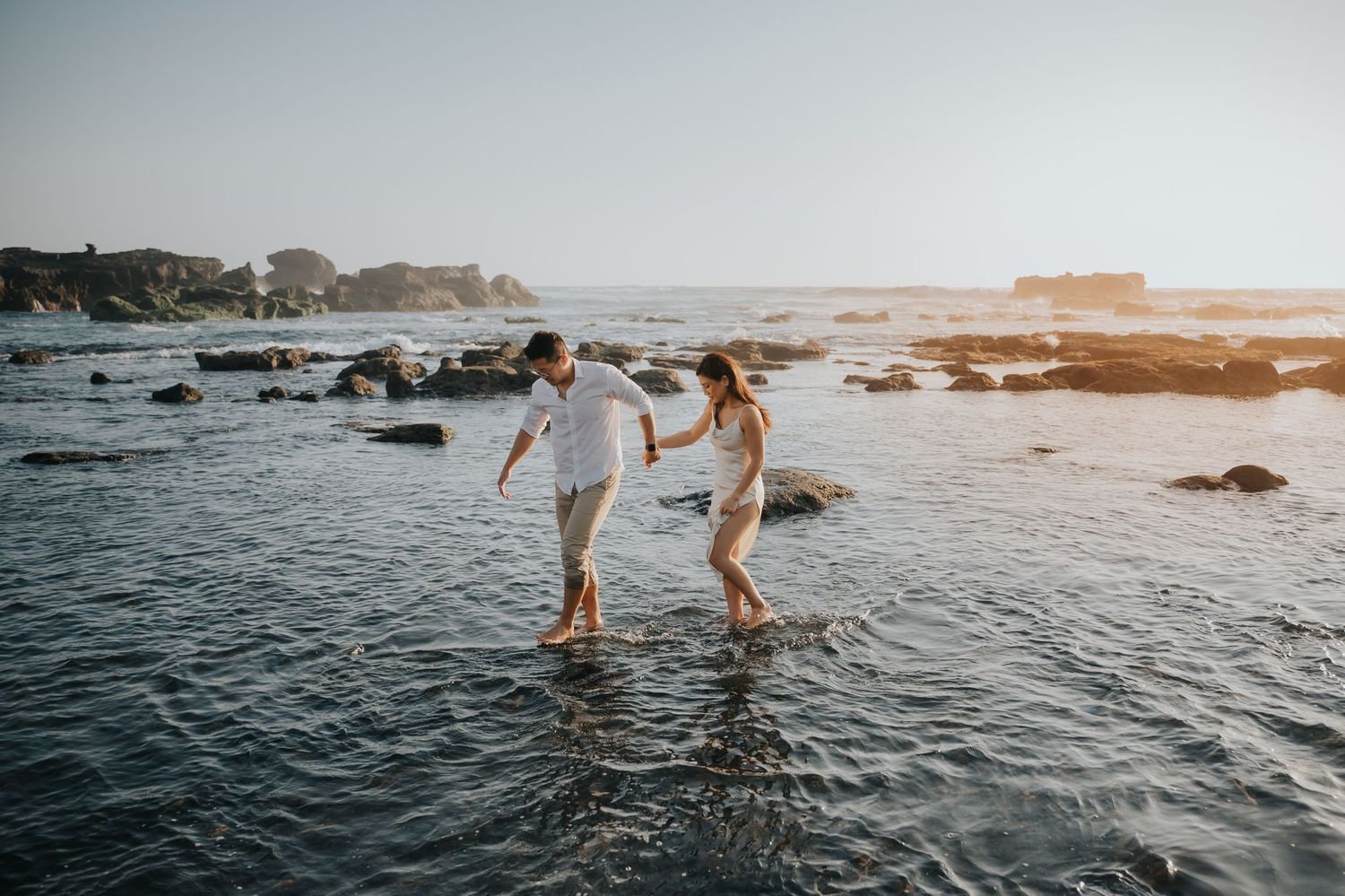 Couple in white outfits holding hands while walking barefoot through shallow ocean water on a rocky beach at sunset.