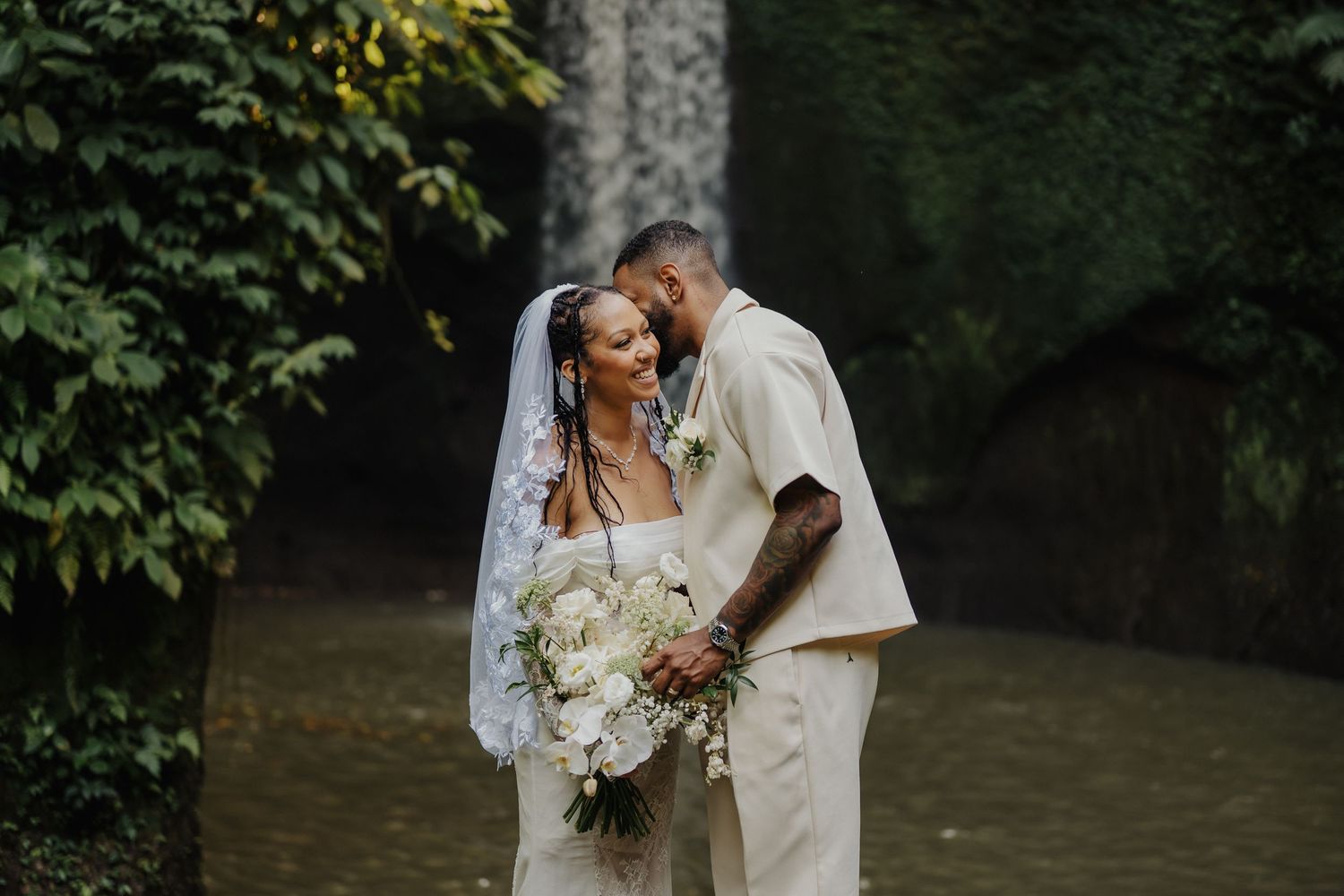 A bride and groom share an intimate moment near a moss-covered waterfall in a lush tropical forest.