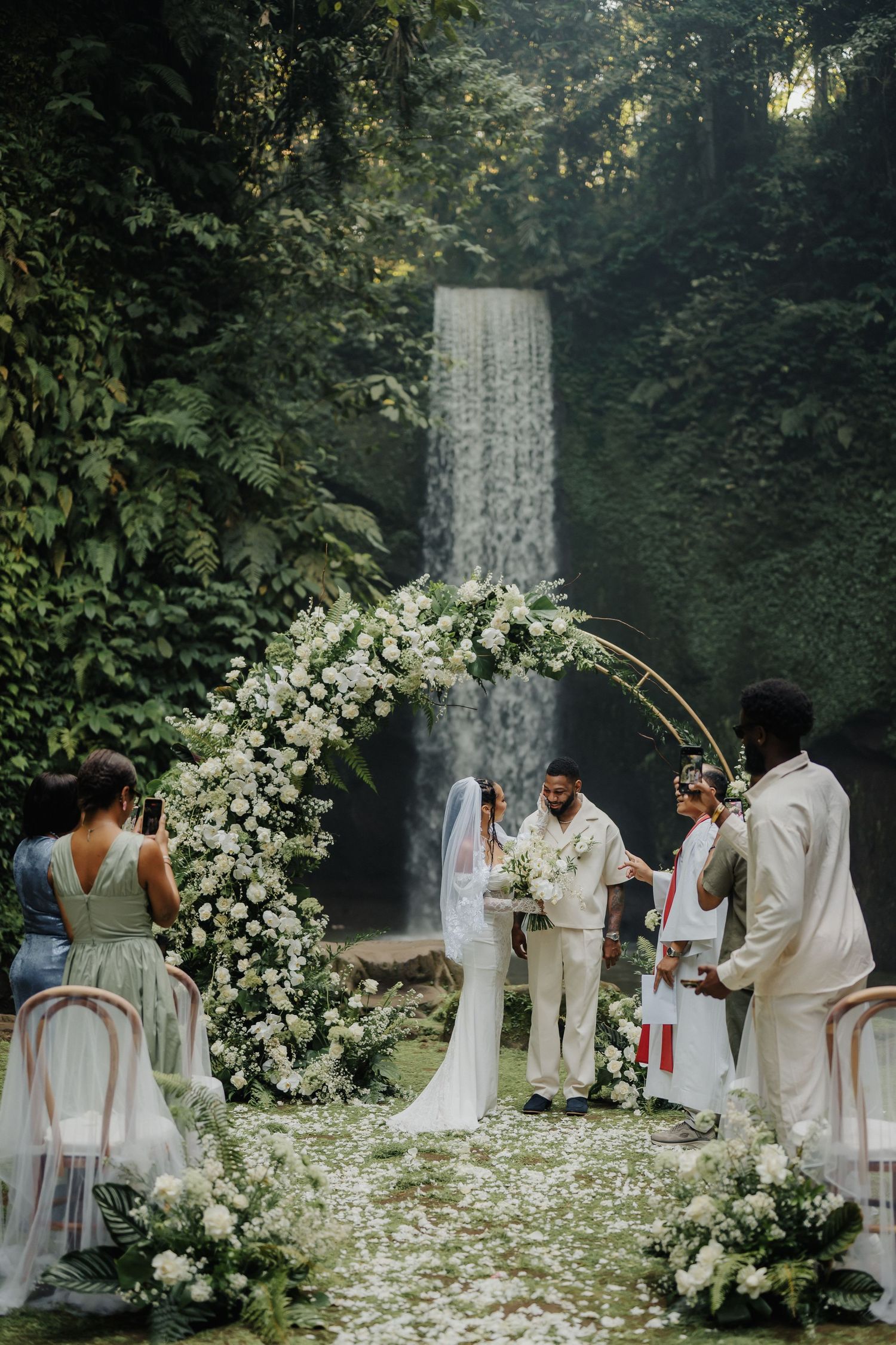 Outdoor wedding ceremony with floral arch and waterfall backdrop surrounded by lush greenery.
