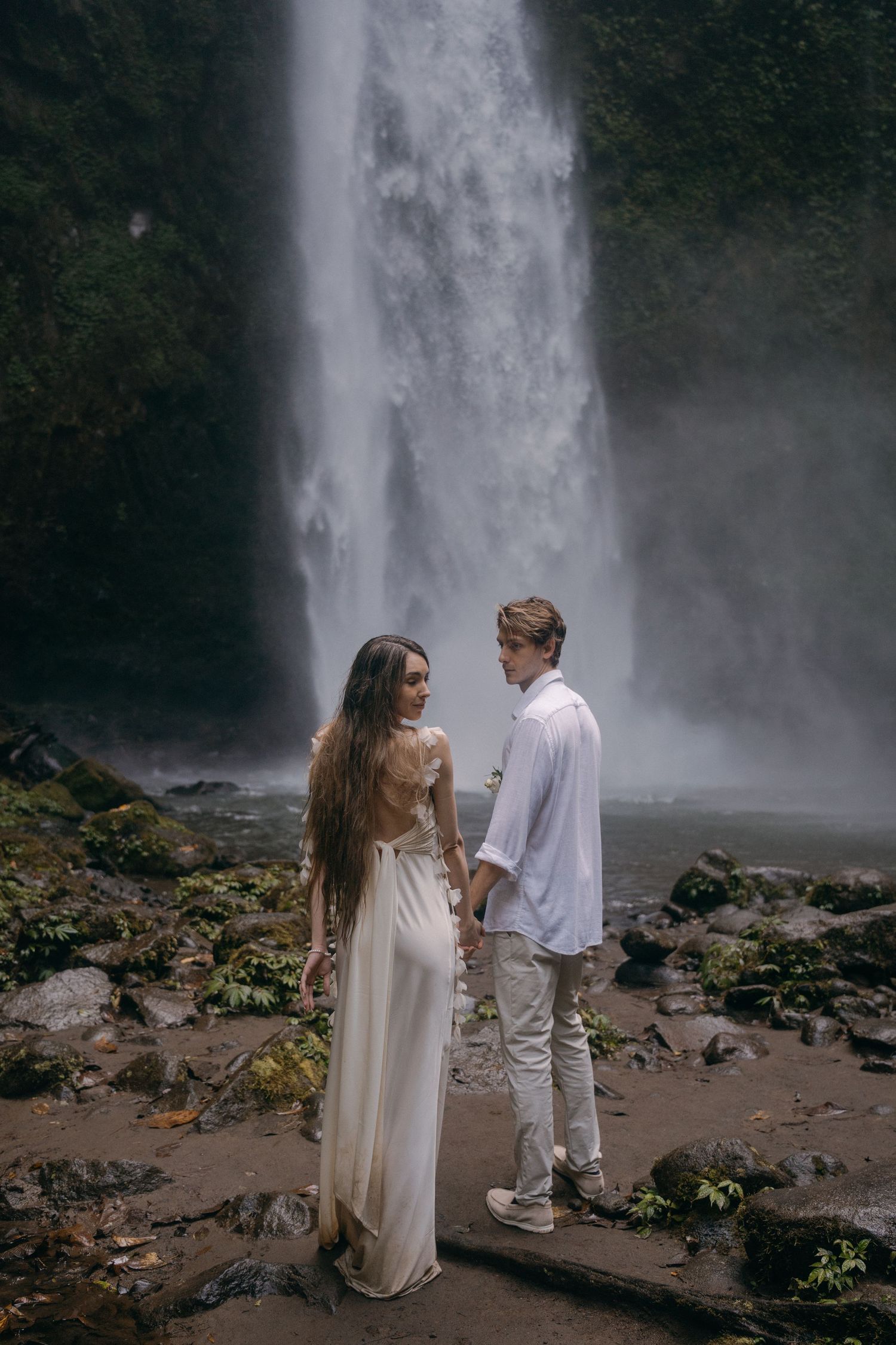 A romantic couple poses near a majestic waterfall on rocky terrain in ethereal white and cream colored outfits.