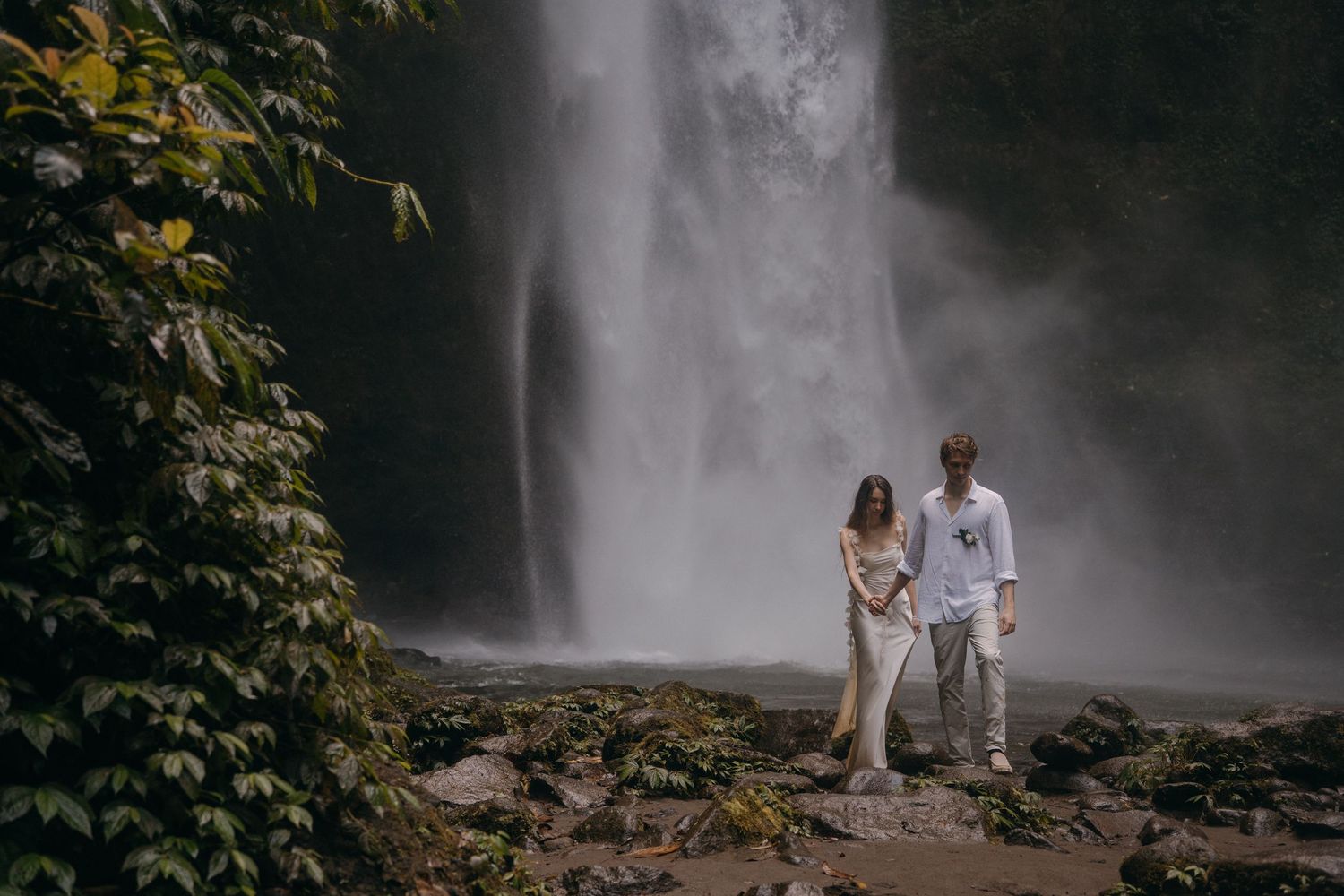 A couple stands together on rocky ground in front of a majestic waterfall surrounded by lush greenery.