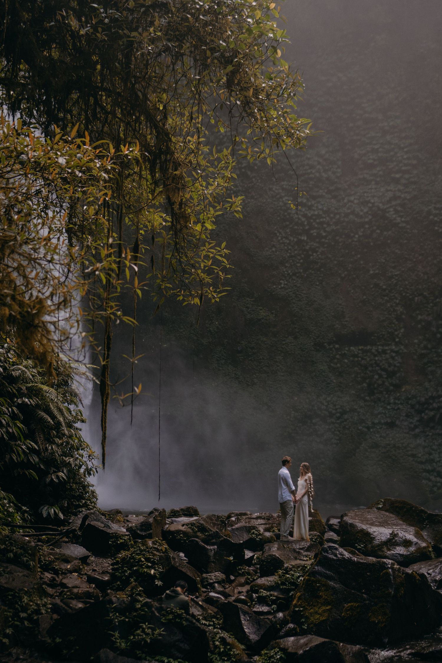 A couple stands in atmospheric fog among rocks and trees at the edge of a misty forest.