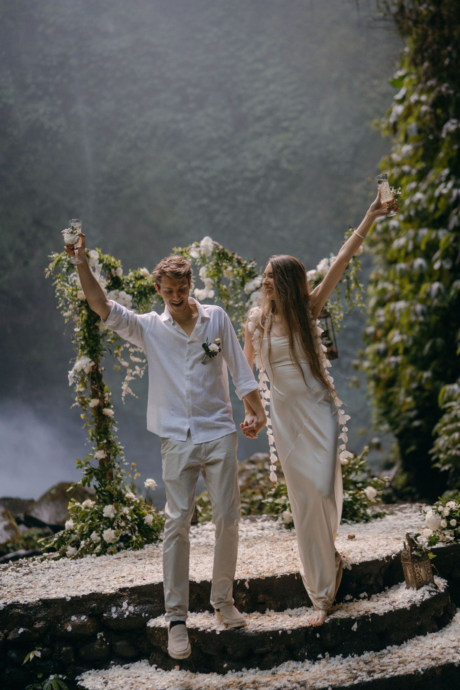 A couple in white outfits dances joyfully on a rocky shore with greenery and mist in the background.