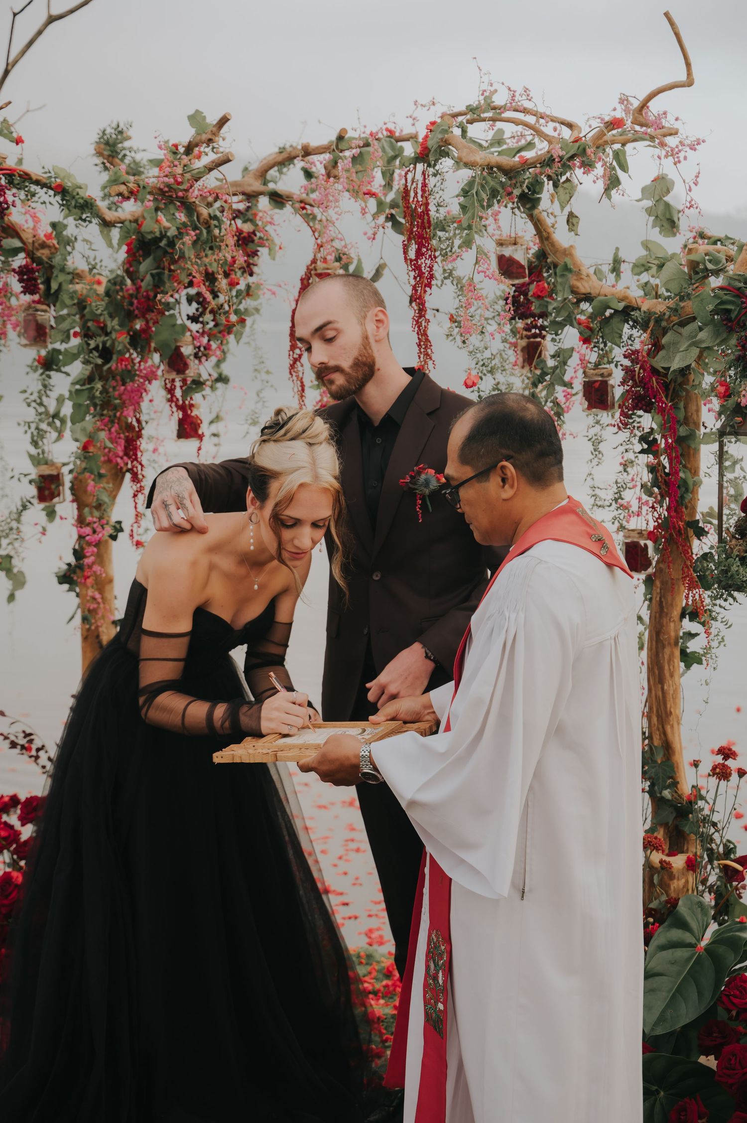 Gothic wedding ceremony with red floral arch and couple in black attire exchanging vows.