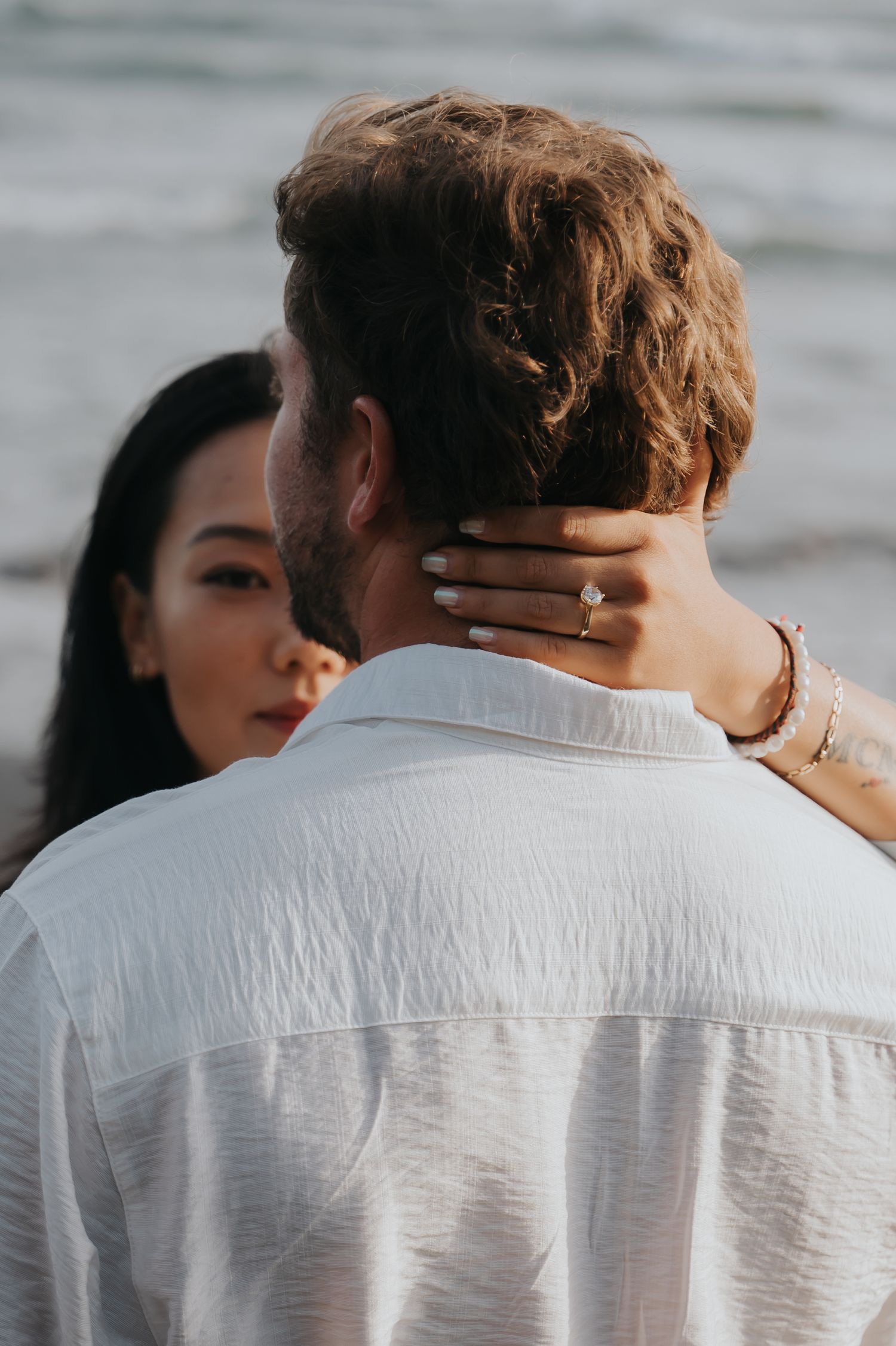 Intimate couple embraces as she gently touches his neck, showing her engagement ring in a soft outdoor setting.
