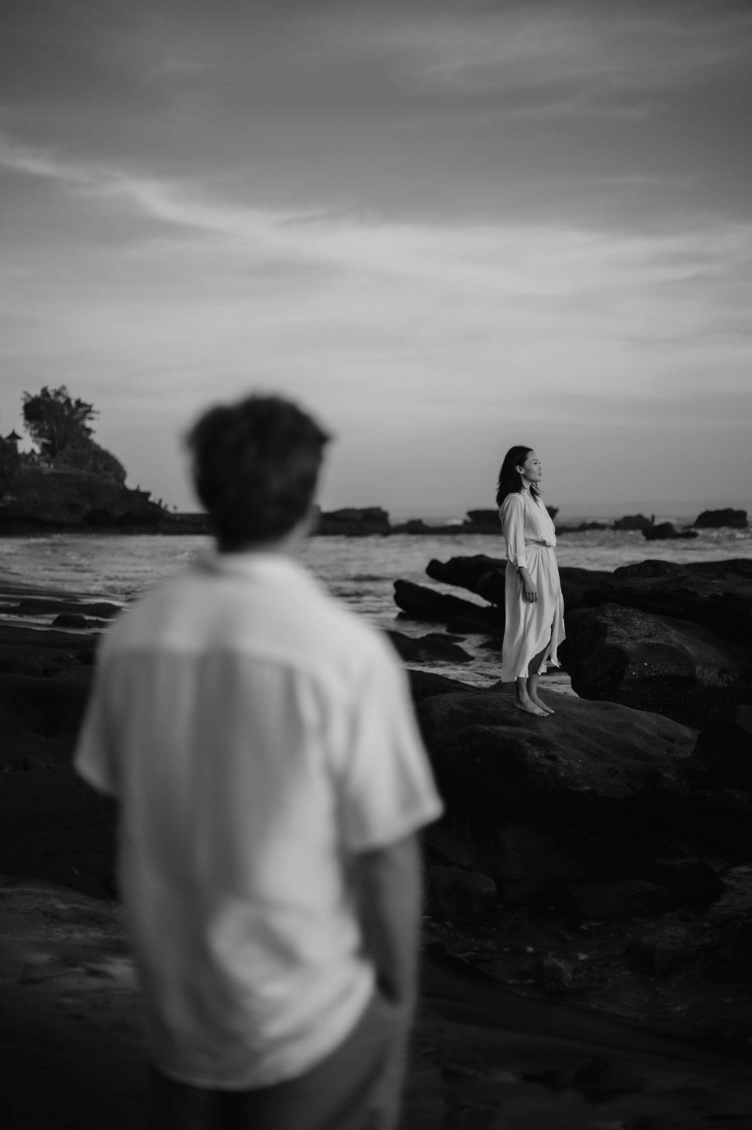 A black and white coastal scene shows two people on rocky shoreline at sunset with dramatic cloudy sky.