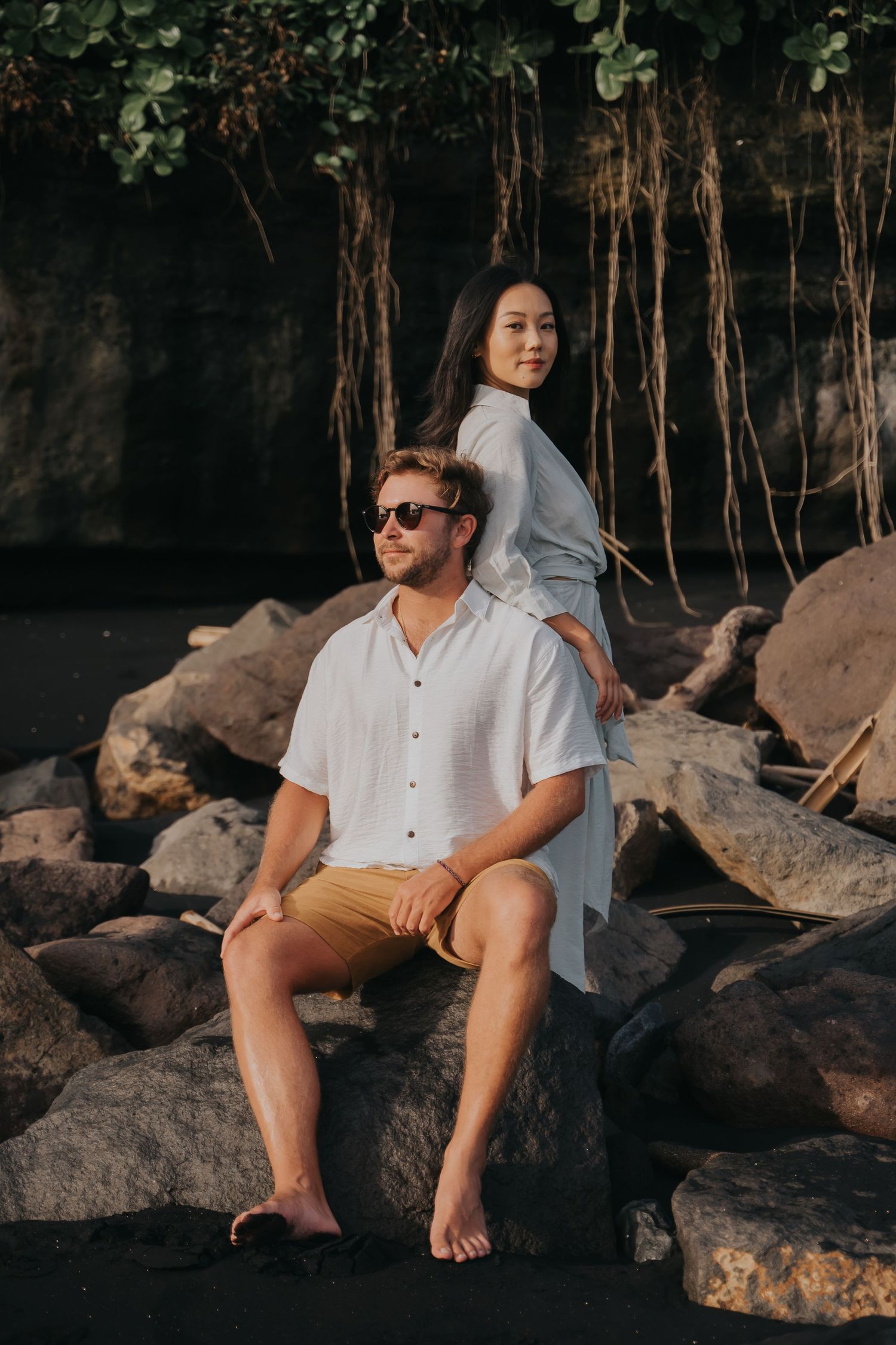 A person in white shirt and shorts sits on coastal rocks at sunset with dramatic shadows and hanging vines in background.