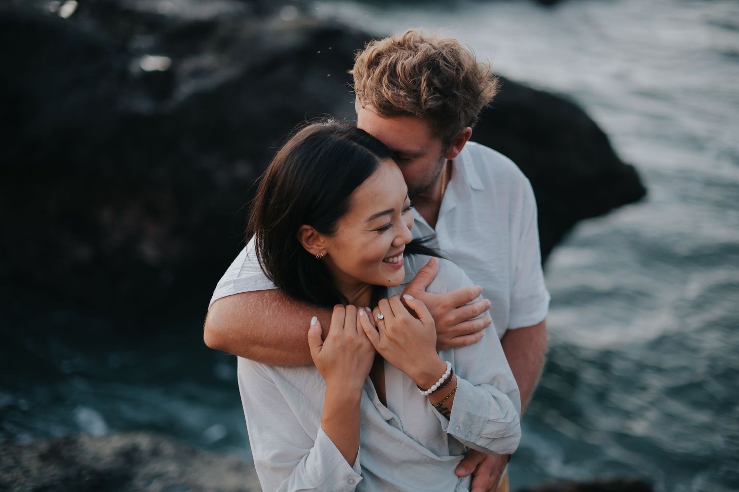 A romantic couple embraces and laughs together during a seaside photoshoot at dusk.