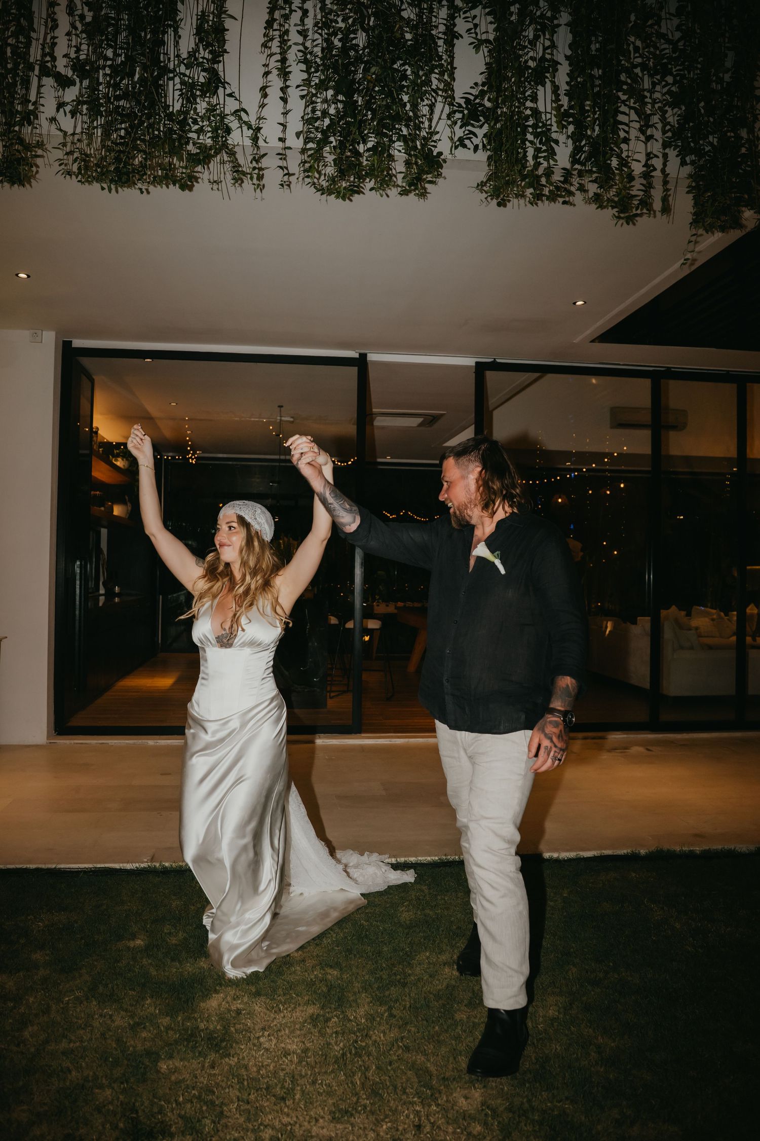 Bride and groom celebrating outdoors at night under hanging greenery with raised hands and smiles.