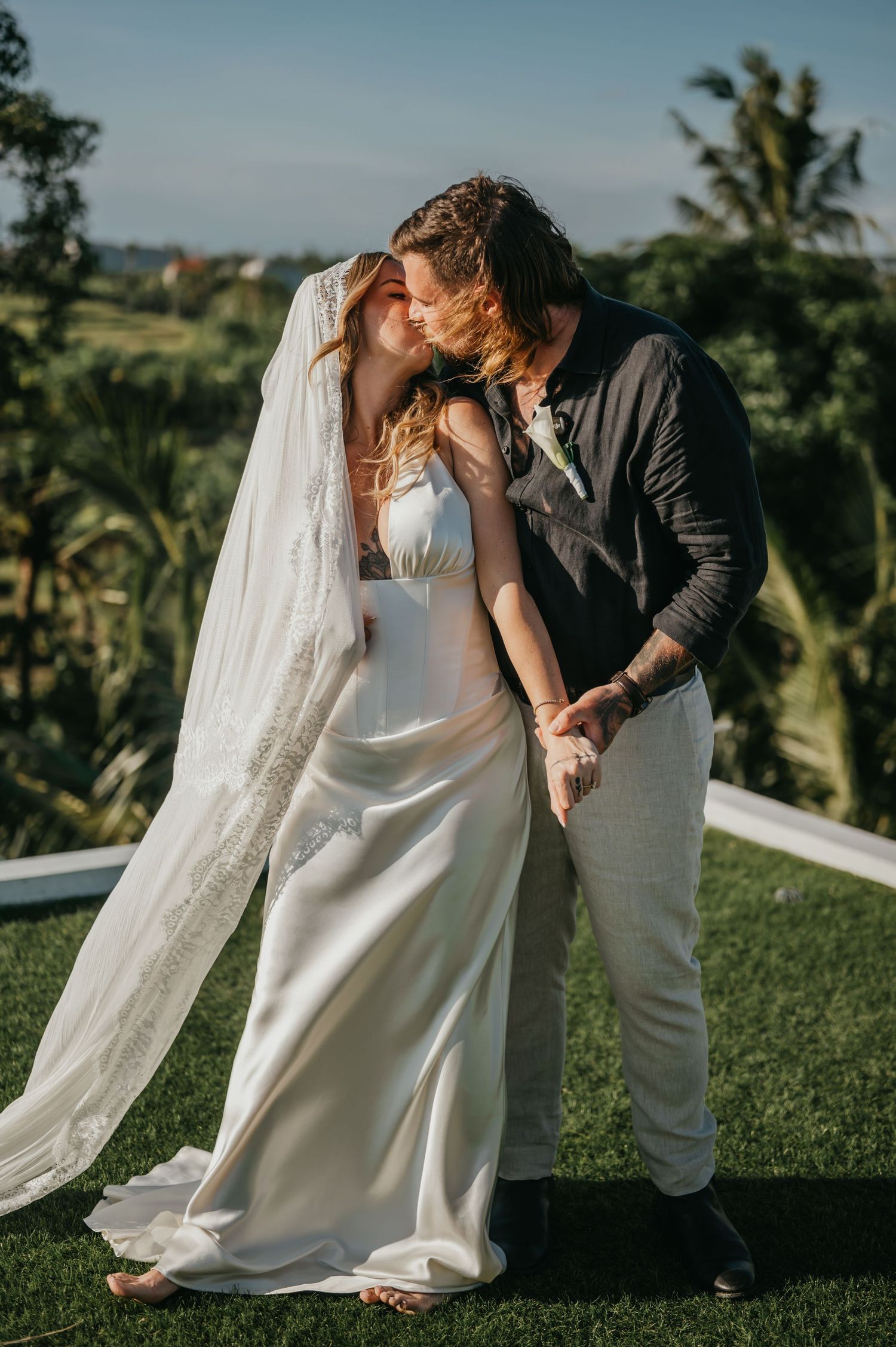 Bride and groom share a romantic kiss on lush green lawn with tropical palm trees in background.