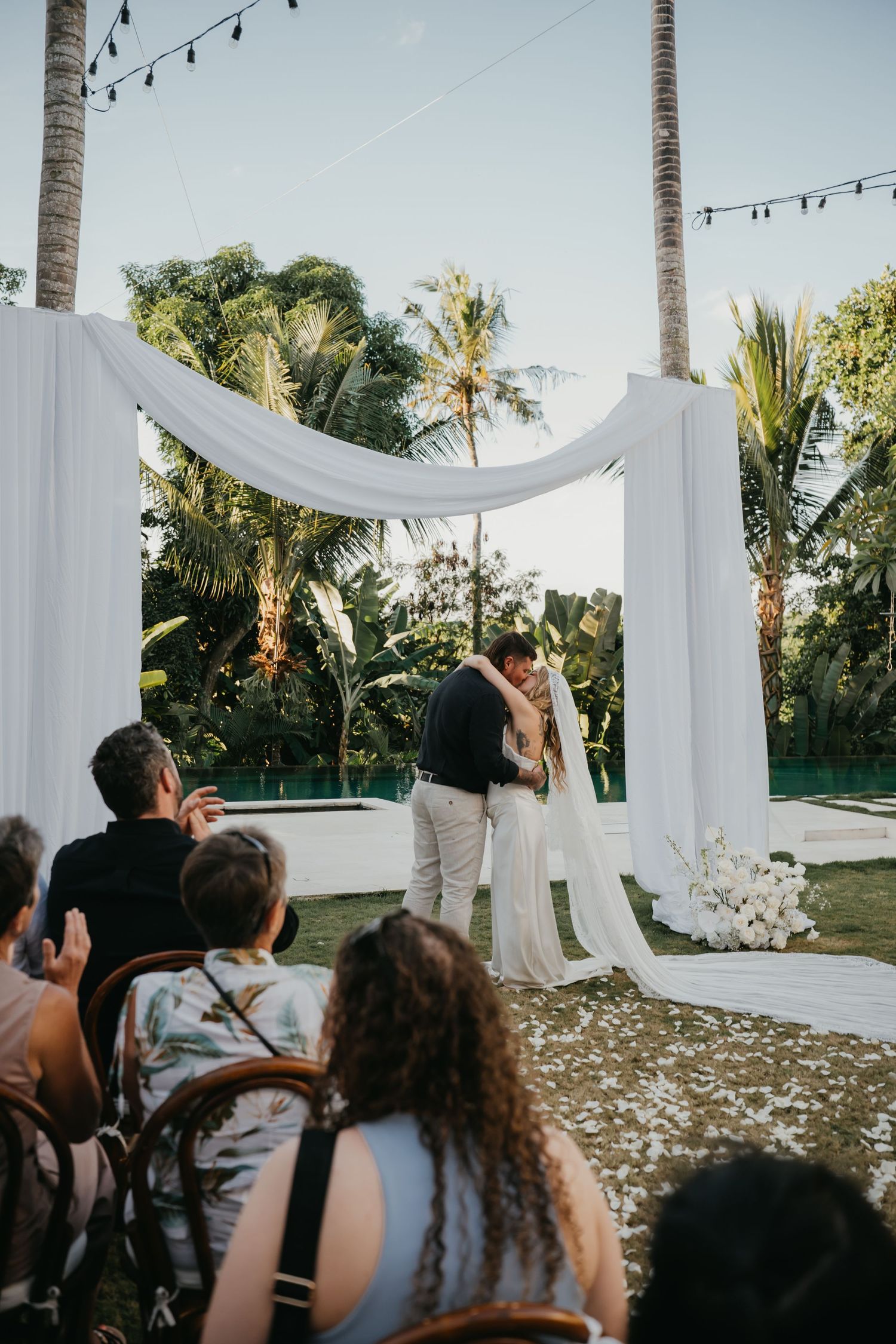 Wedding couple shares first kiss under elegant white draped arch while guests watch at tropical outdoor ceremony.