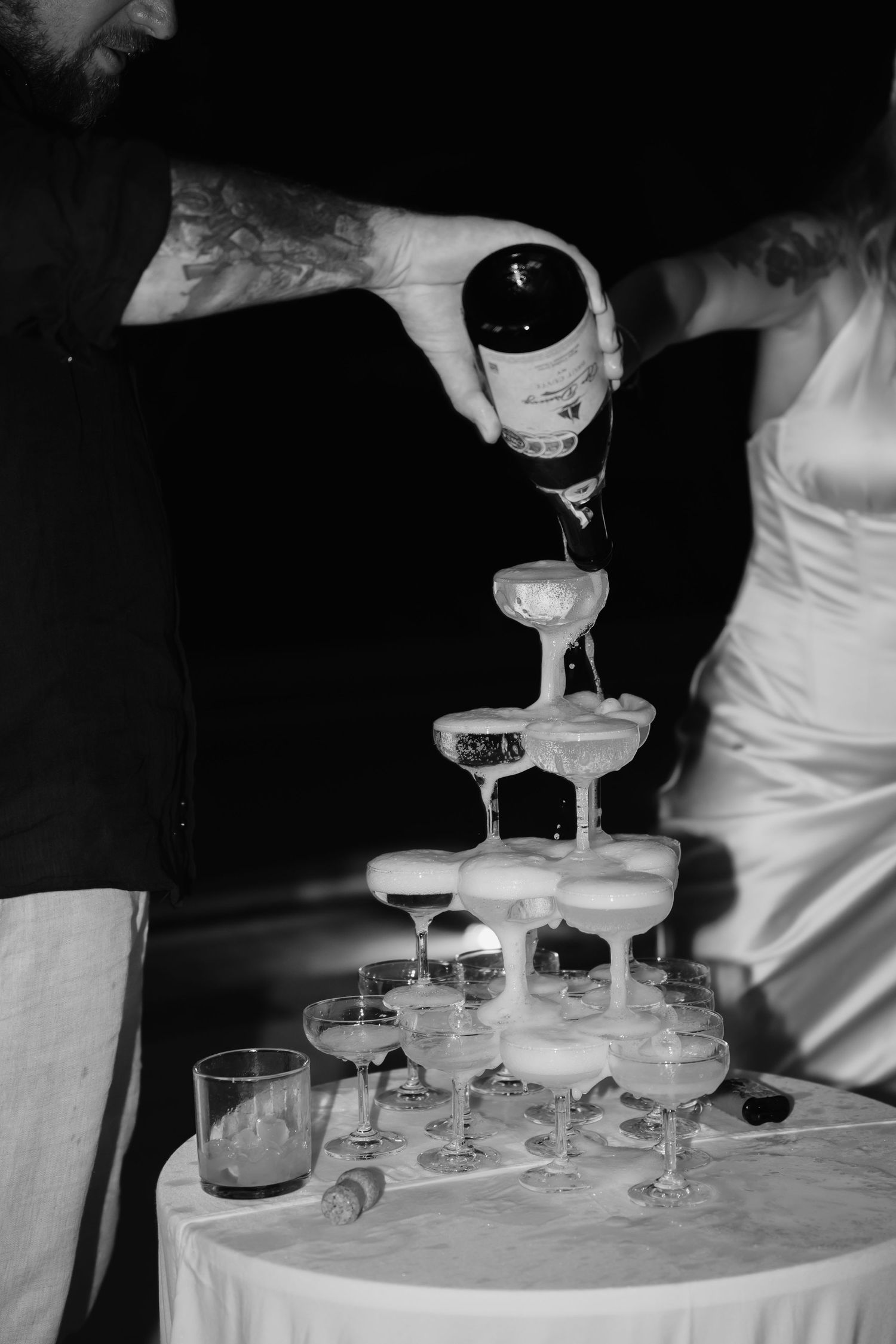 Tattooed groom pours champagne over tiered coupe glass tower beside bride in white gown in black and white photo.