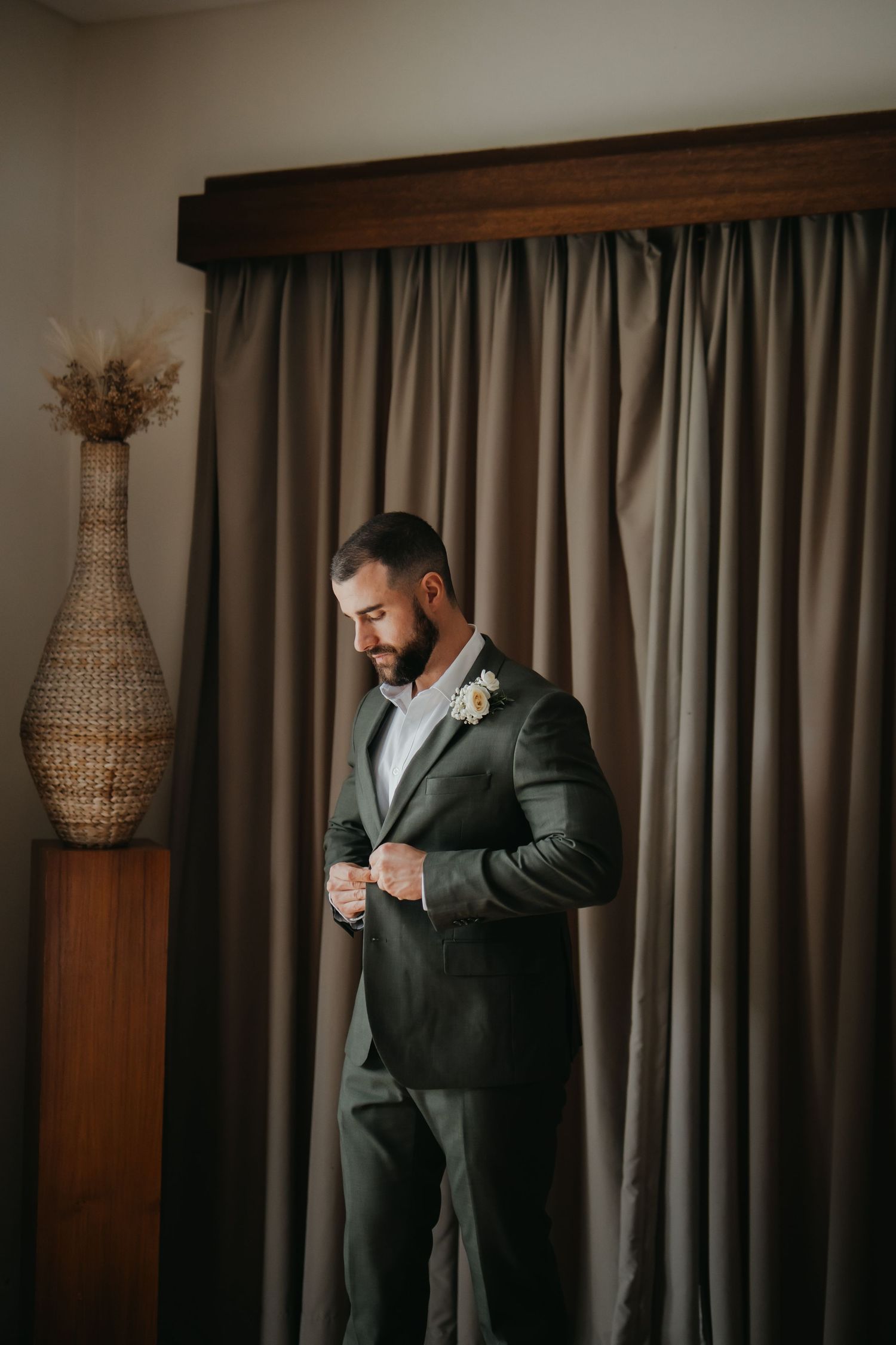 A handsome groom in a dark suit adjusts his jacket button while standing before gray curtains.